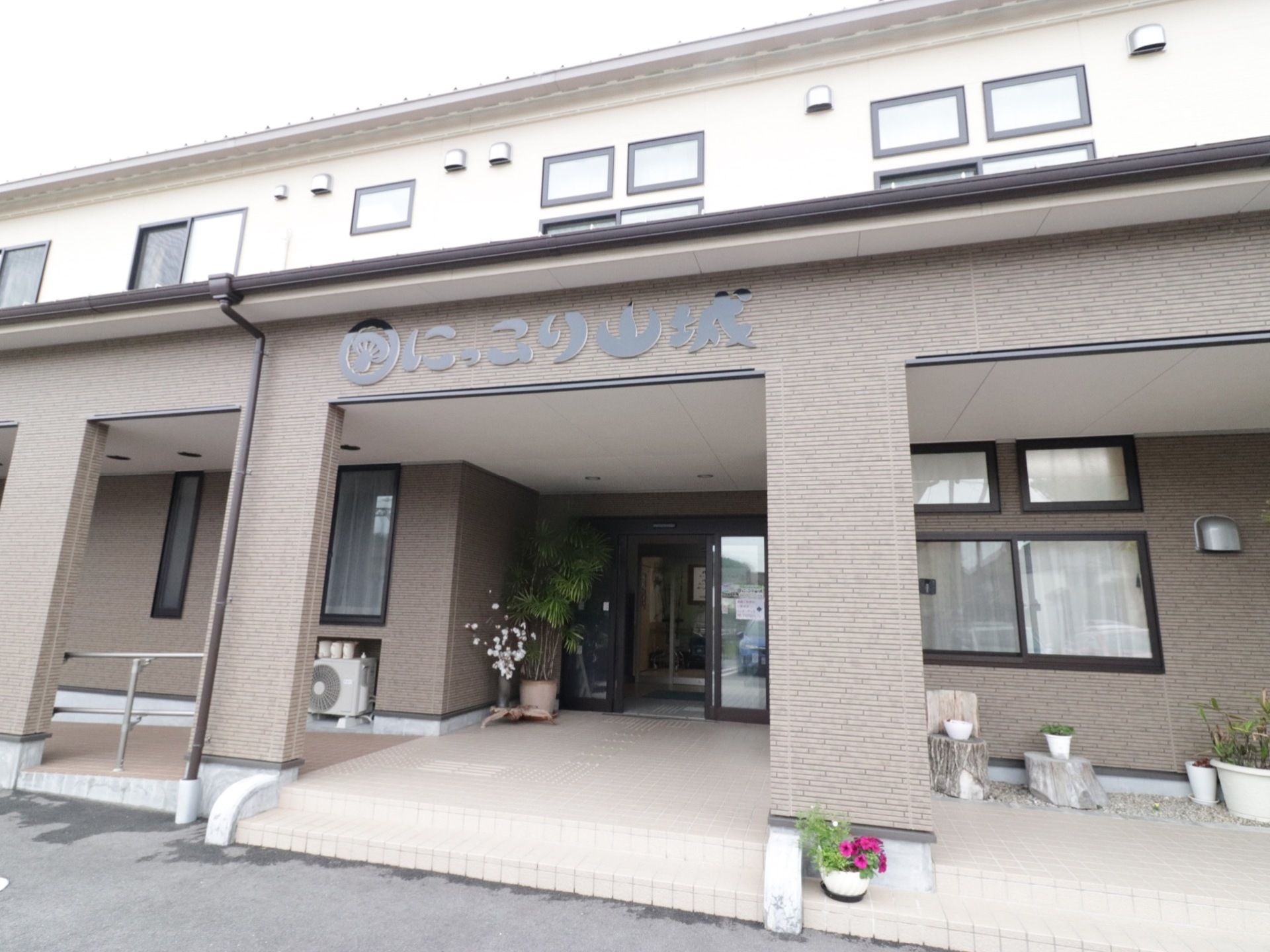 ２階建てで横長の建物がある。手すりのついたスロープと、階段の両方がある。玄関周りは鉢植えが飾られている。