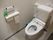 自動洗浄器付きの洋式トイレである。便座のフタは外してあり、後ろの棚に予備のトイレットペーパーが置いてある。