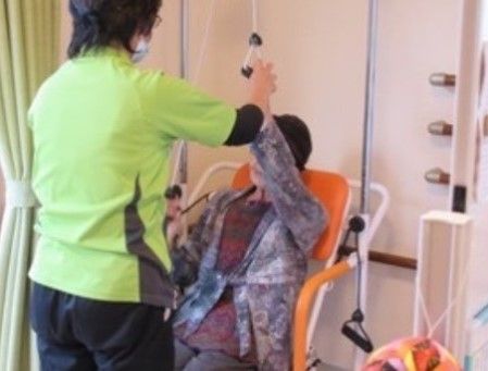 オレンジの椅子に座り腕のリハビリをする入居者様とサポートするスタッフ