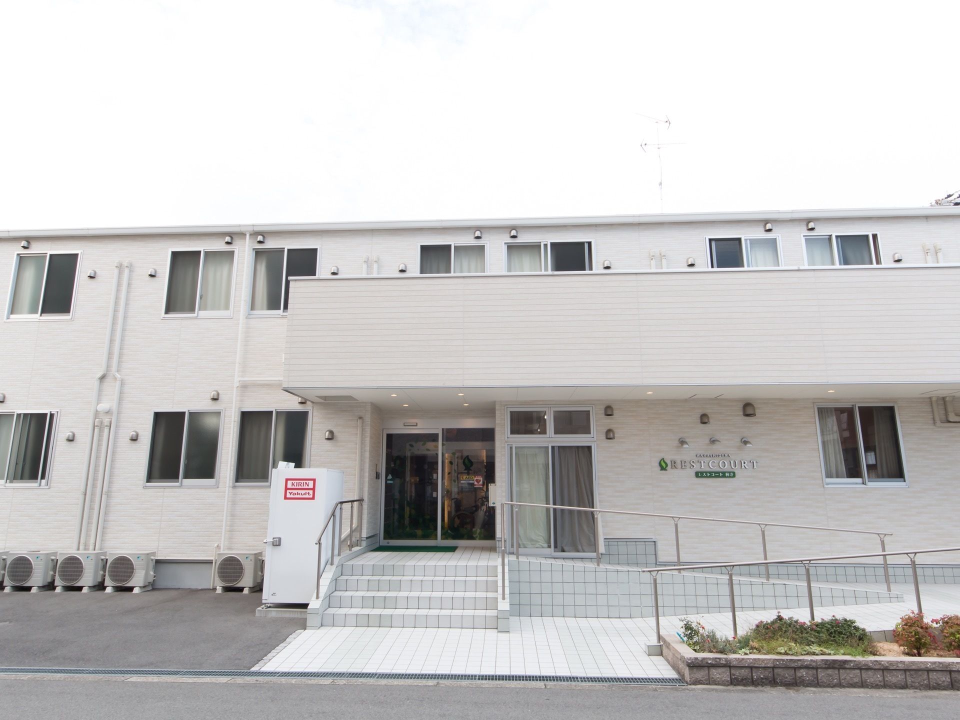 白い外壁の2階建ての建物があり、2階にはベランダが設置されている。施設前には自動販売機が置かれている。