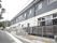 道路に沿って2階建ての建物がある。建物と道路の間に芝生帯がある。道路との境にグレー色のフェンスがある。