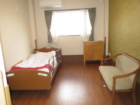 居室には介護用ベッドと2人掛けチェアーを置いてある。壁の一部がモスグリーンの壁紙になっていて、おしゃれで落ち着きのある部屋になっている。