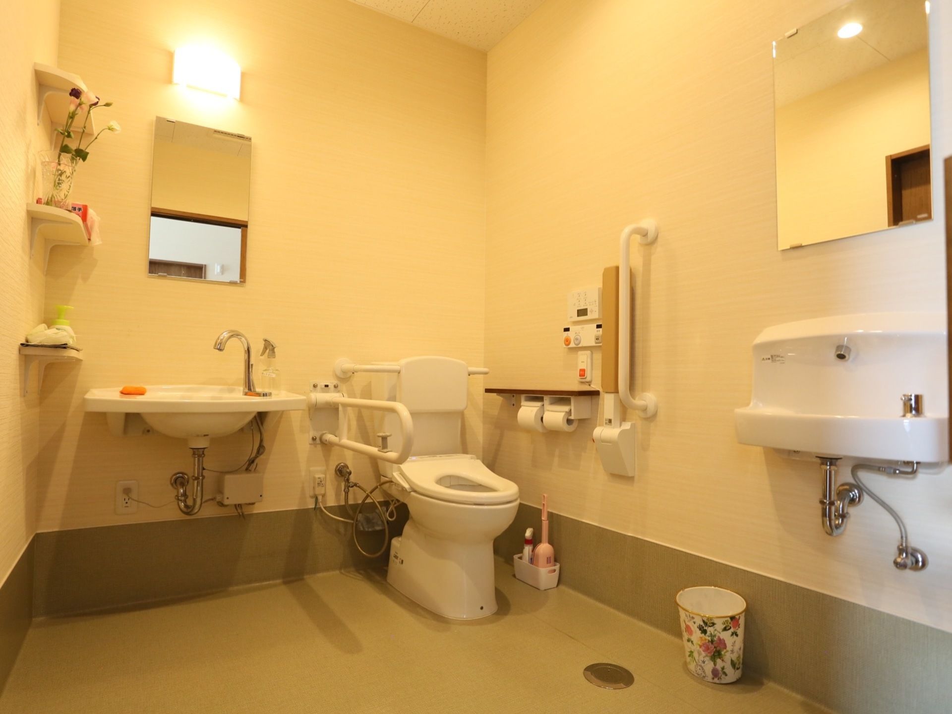 背もたれが設置された洋式トイレと洗面台が２か所ある。洗面台のそれぞれ壁に鏡が取りつけられている。床にゴミ箱が置いてある。
