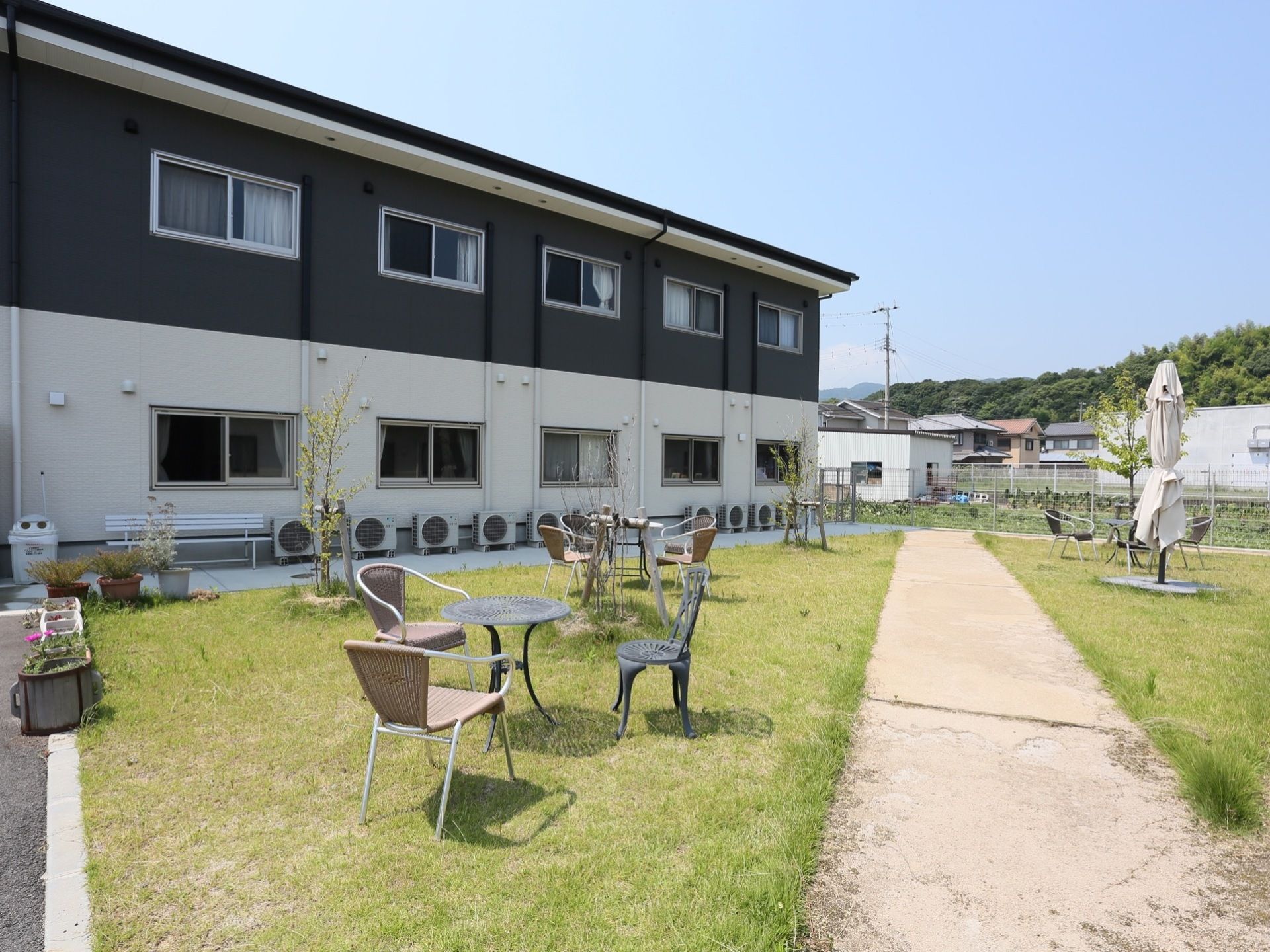 芝生が敷かれた庭にテーブルや椅子が置いてある。白と黒の２階建ての建物が建っている。建物に沿ってエアコンの室外機が設置されている。