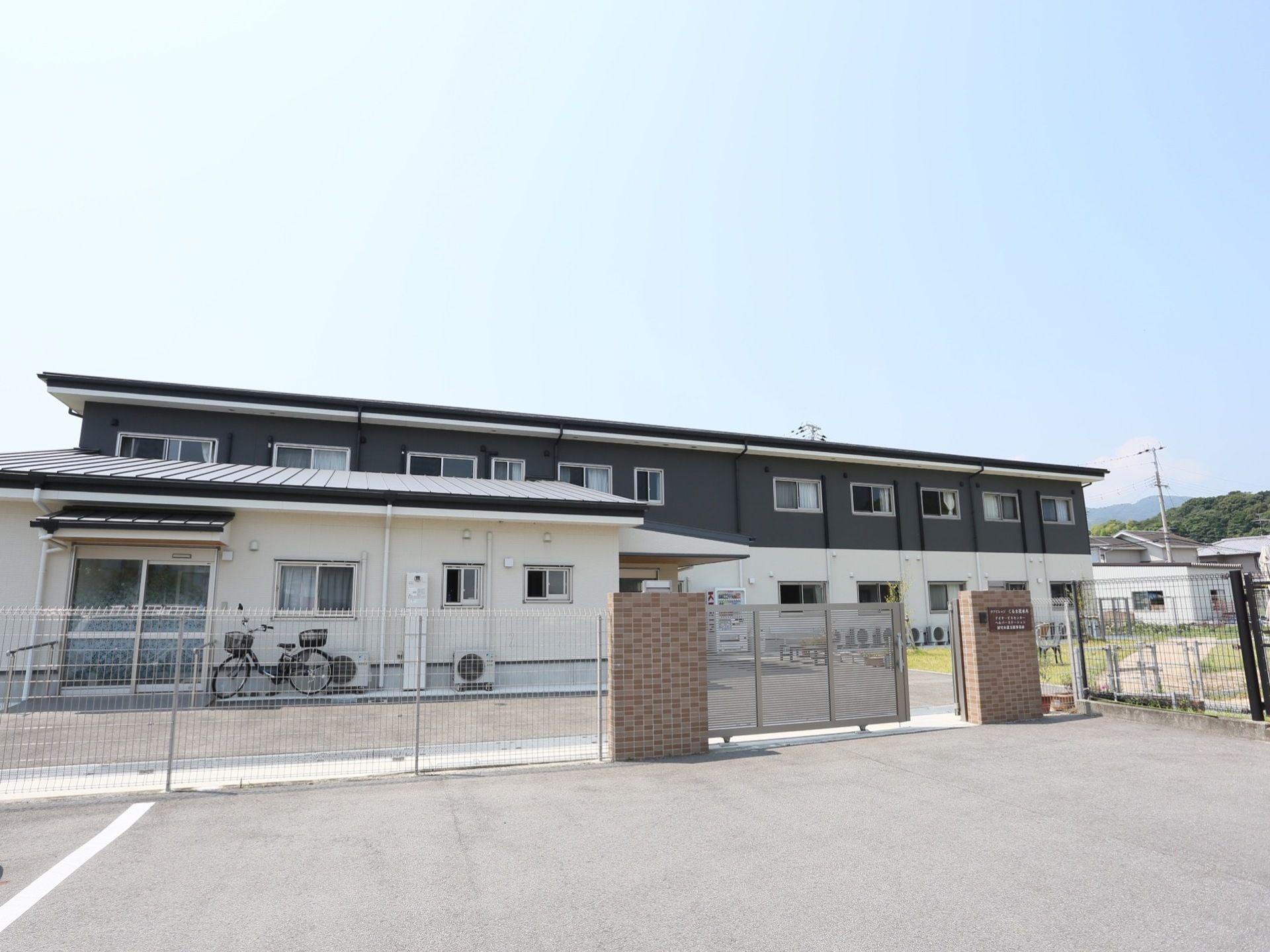 １階が白い壁で２階が濃紺の壁の２階建ての建物がある。駐車場と敷地はフェンスで仕切られ、入り口はスライドタイプの門扉が設置されている。