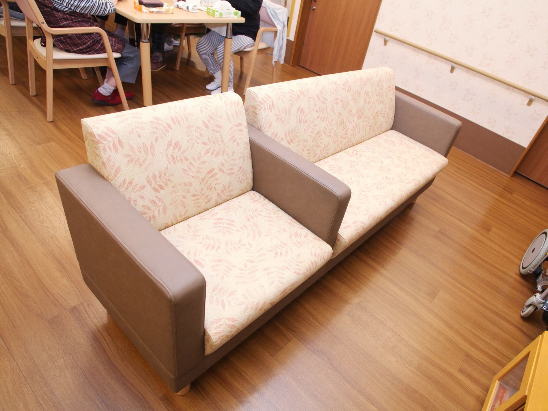 淡いピンク色のソファーが置かれている。壁も淡いピンク色になっており、手すりが付いている。後ろにはテーブルと椅子があり、人が座っている。