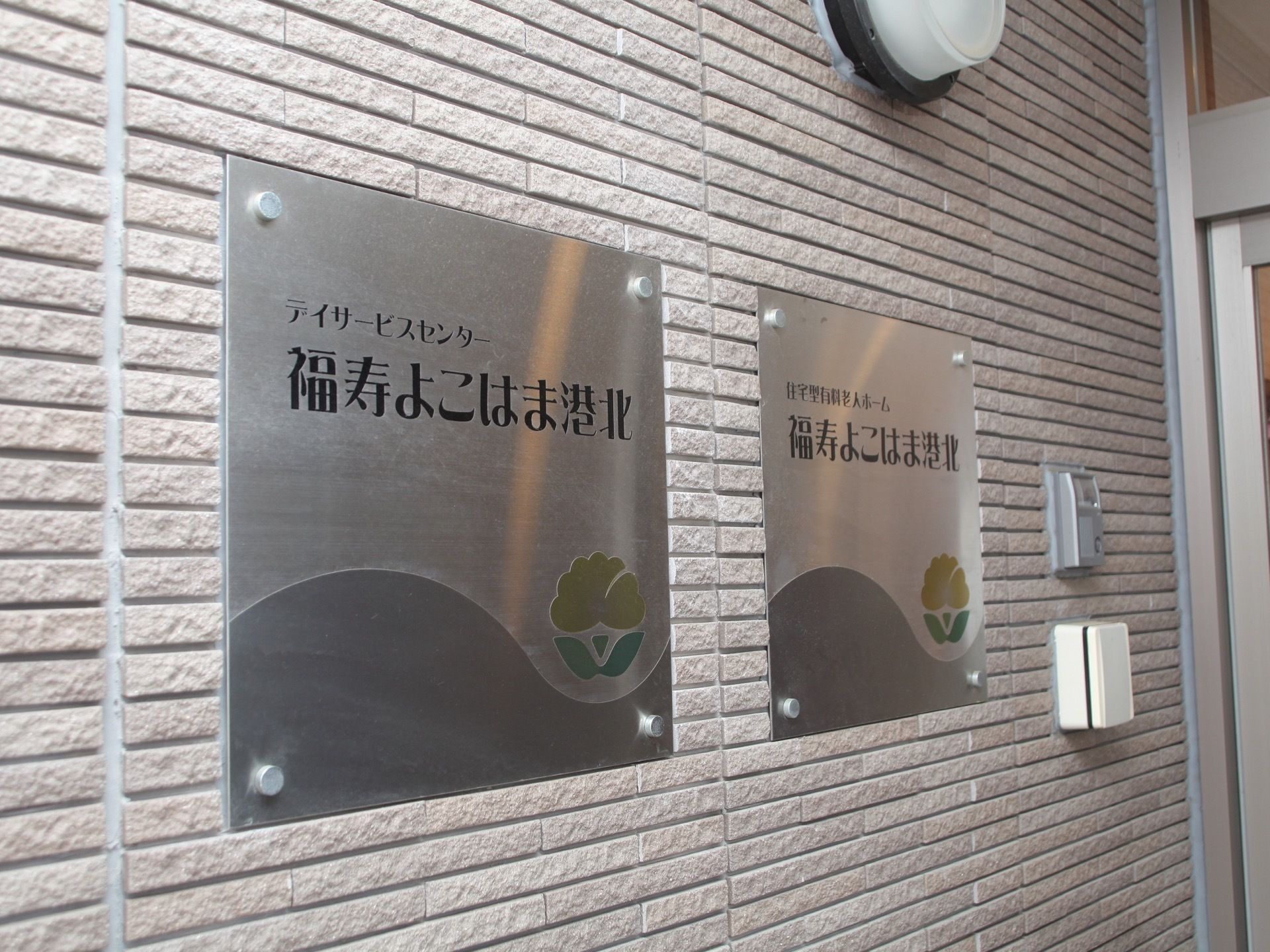 金属製のプレートに施設名が書かれた看板が２つ並んでいる。レンガ調の壁にはインターホンや、照明が設置されている。