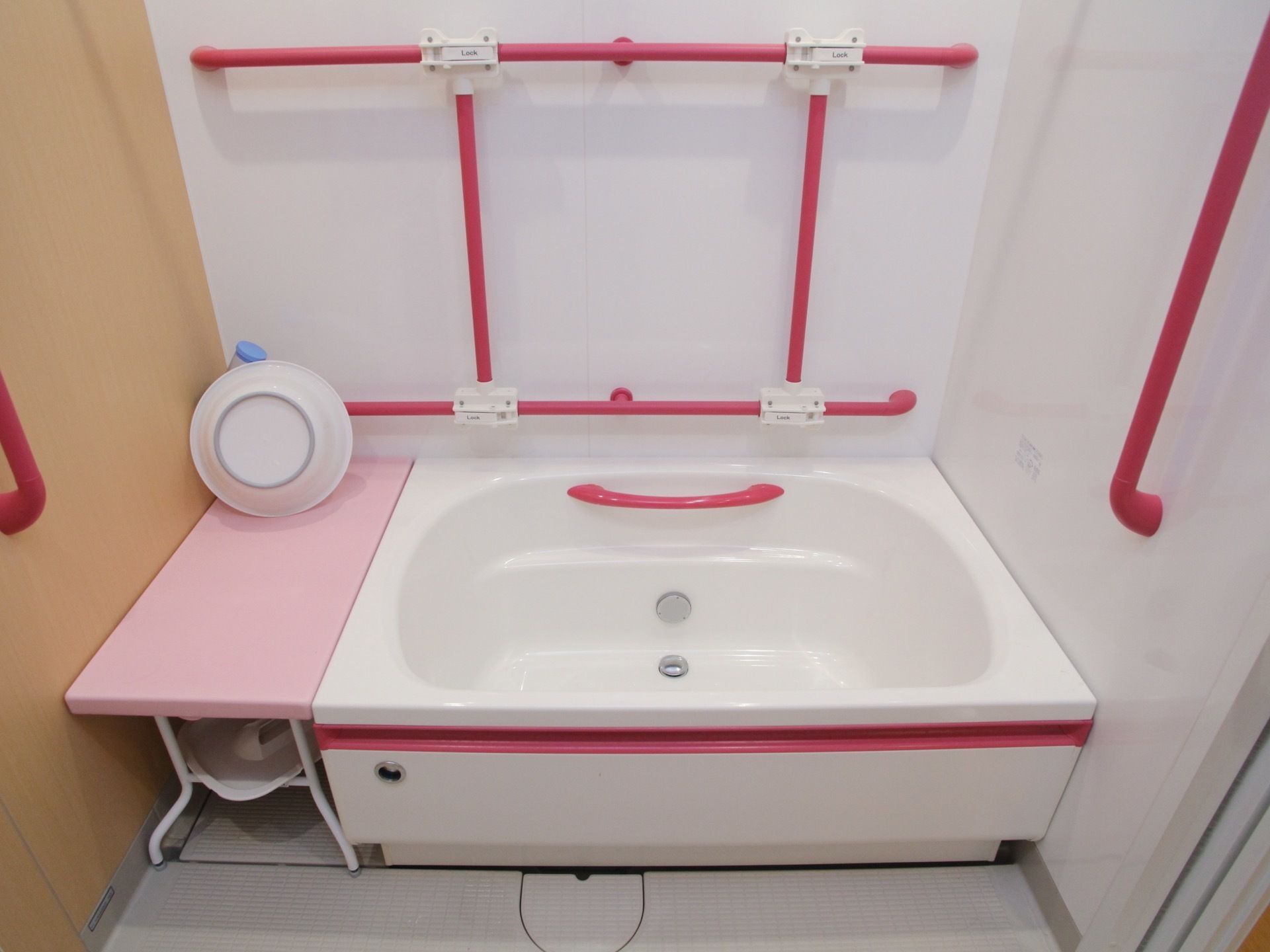 壁や浴槽は白くて、赤い手すりが付いている。ピンクの台には洗面器が置かれ、そこに腰かけることもできる。