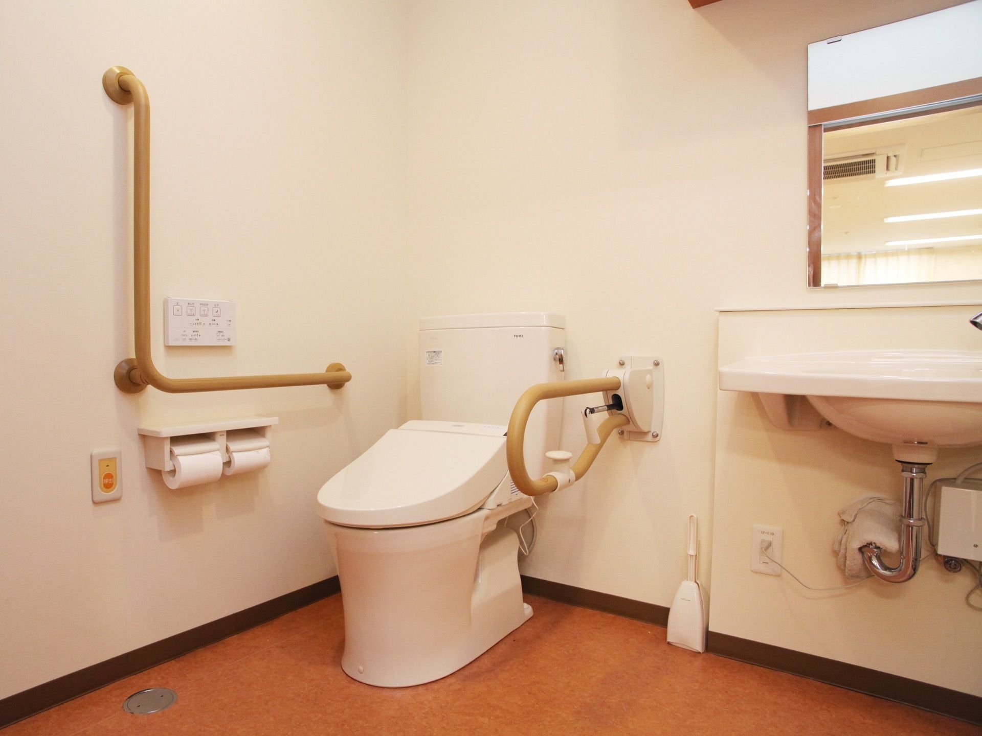 温水洗浄便座が設置され、壁には操作ボタンや手すり、トイレットペーパー、呼び出しボタンが付いている。明るく清潔感のある雰囲気がある。