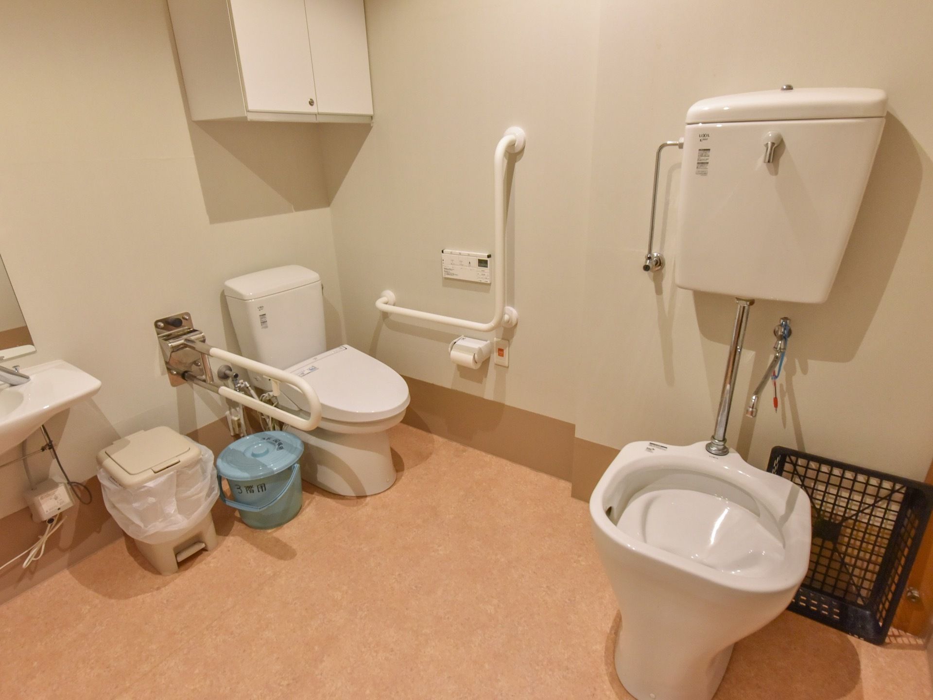 広々としたトイレには、便座が2つと簡易洗面台が設置されている。室内には、青いバケツと大きなゴミ箱が置かれている。