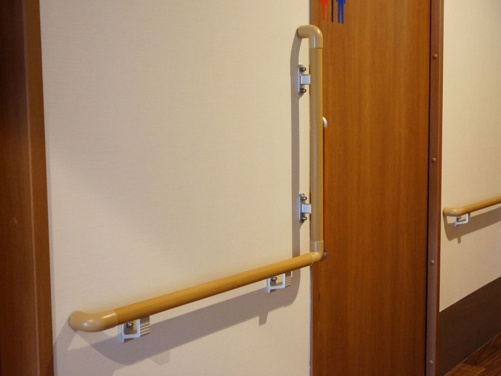 白色の廊下の壁に、茶色の手すりが設置されている。共用トイレのドアがあり、その横は掴まりやすいようL字型になっている。