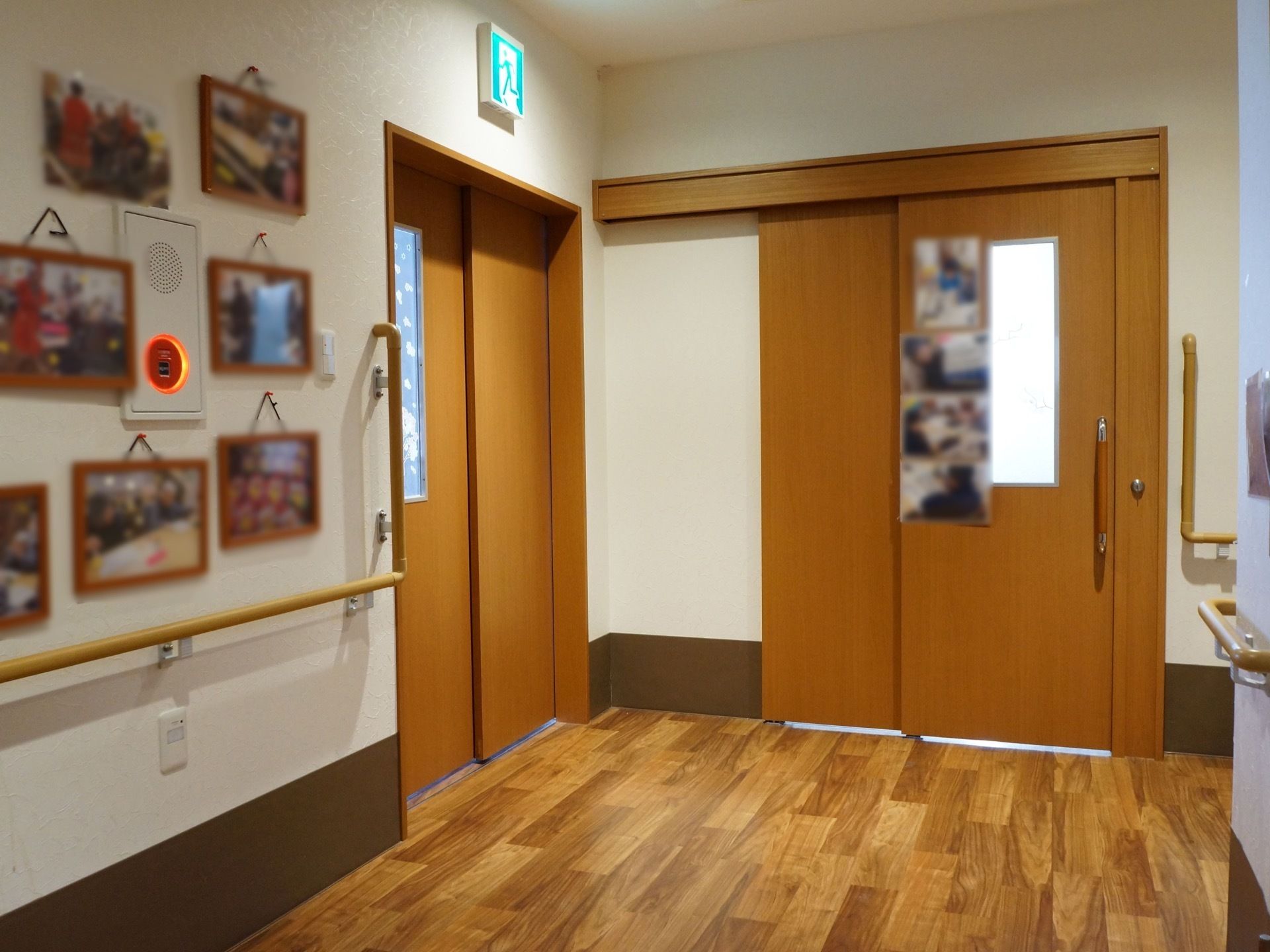 フローリングの床に白い壁の廊下で、茶色のドアが二枚あり、片方は非常口である。ドアの横は縦型にも手すりが付いている。