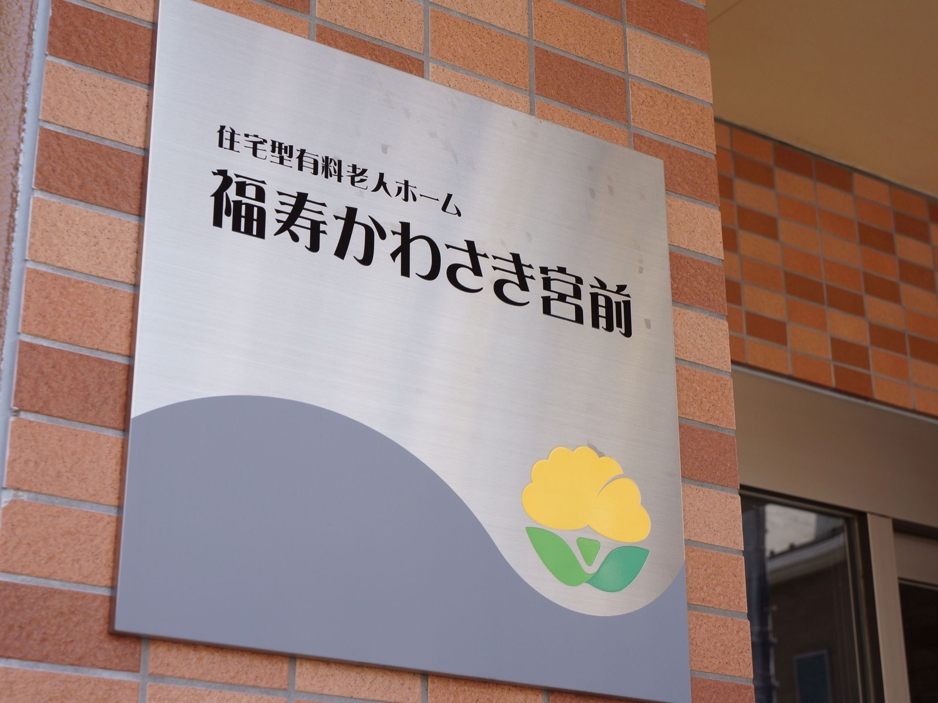 玄関のレンガ風の外壁に、グレーの正方形の看板が付いている。施設の名称とシンボルマークが描かれている。
