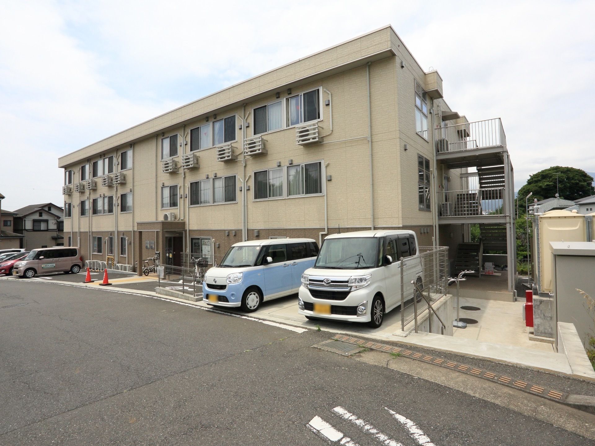 玄関を出たところに駐車場がある。複数の車が停められ、来訪者様が来られるときも安心である。目の前は道路が走っている。
