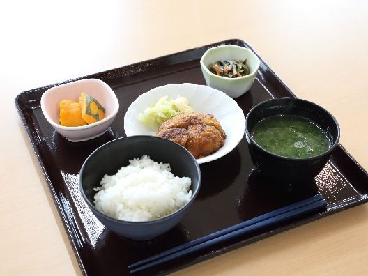 野菜が添えられたメンチカツや白いご飯、みそ汁、かぼちゃの煮つけなどの小鉢が並んだ黒いトレー。