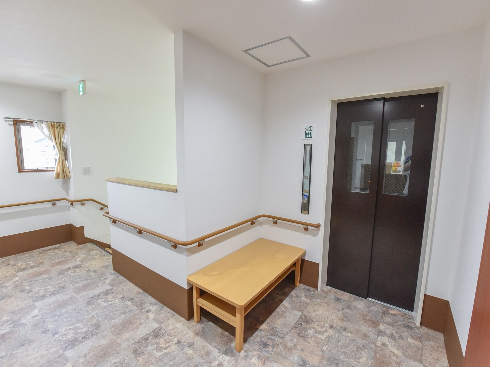 窓がついた黒い扉のエレベーターがある。手すりが設置された壁の前に長椅子が置いてあり、小窓にカーテンが掛けられている。