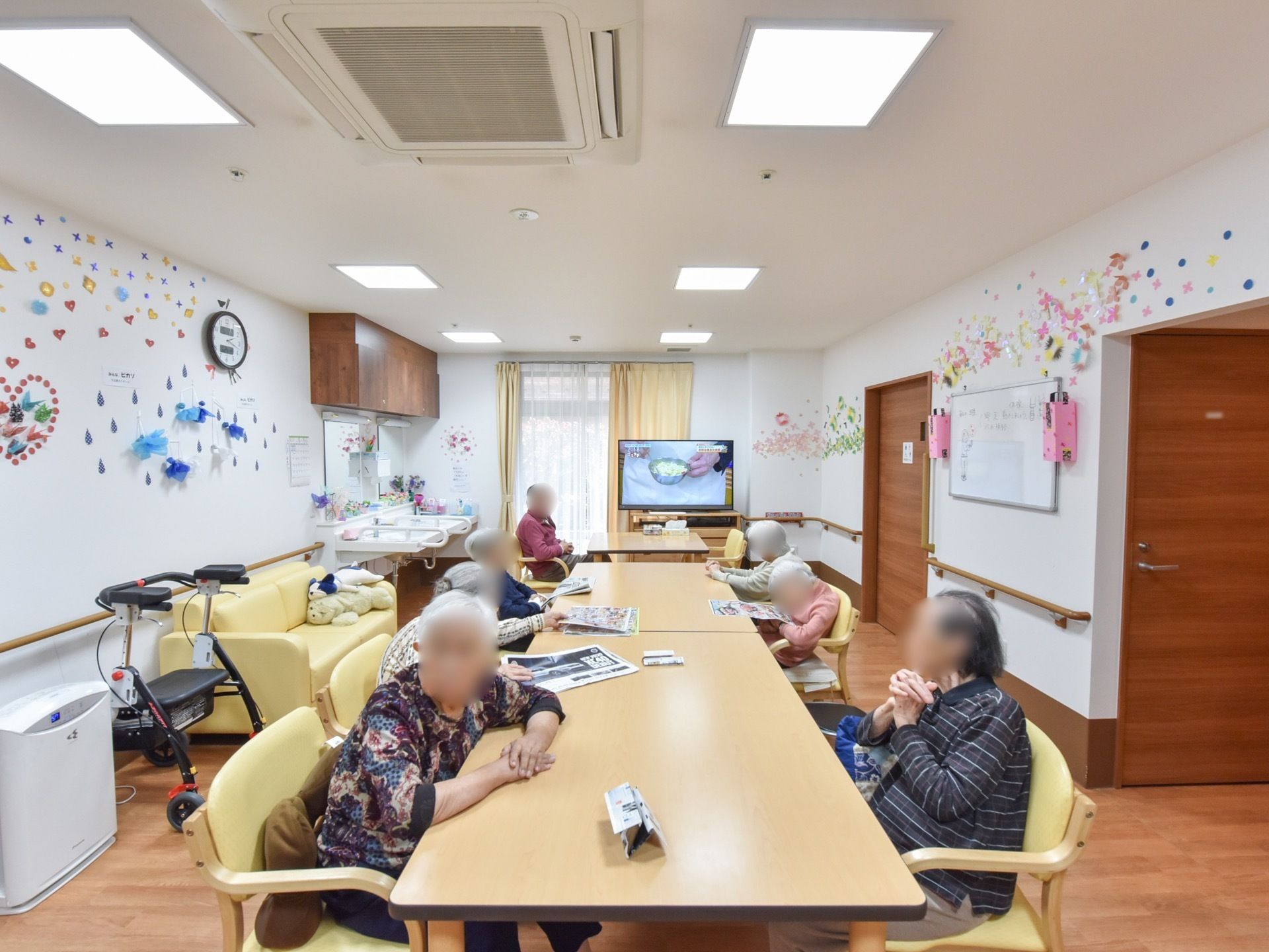 部屋の中央にテーブルが配置され、両側に入居者様が腰を掛けている。壁にたくさんの飾り付けがされ、天井の照明が点灯している。