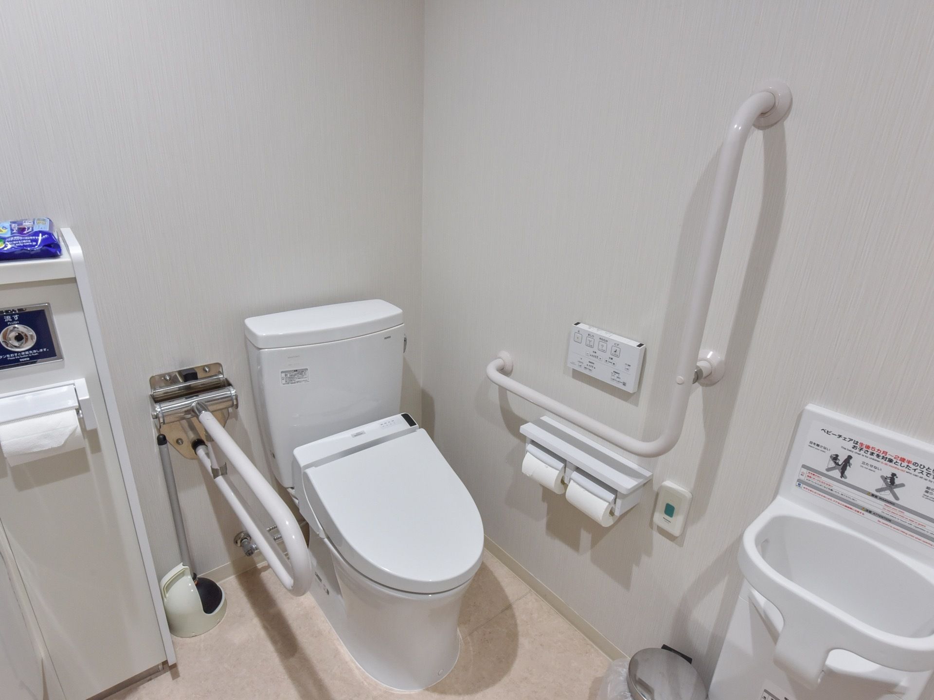 タンク付きの白い洋式トイレに可動式の手すりが取り付けられている。壁に手すりと温水洗浄便座のリモコンとペーパーホルダーが設置されている。