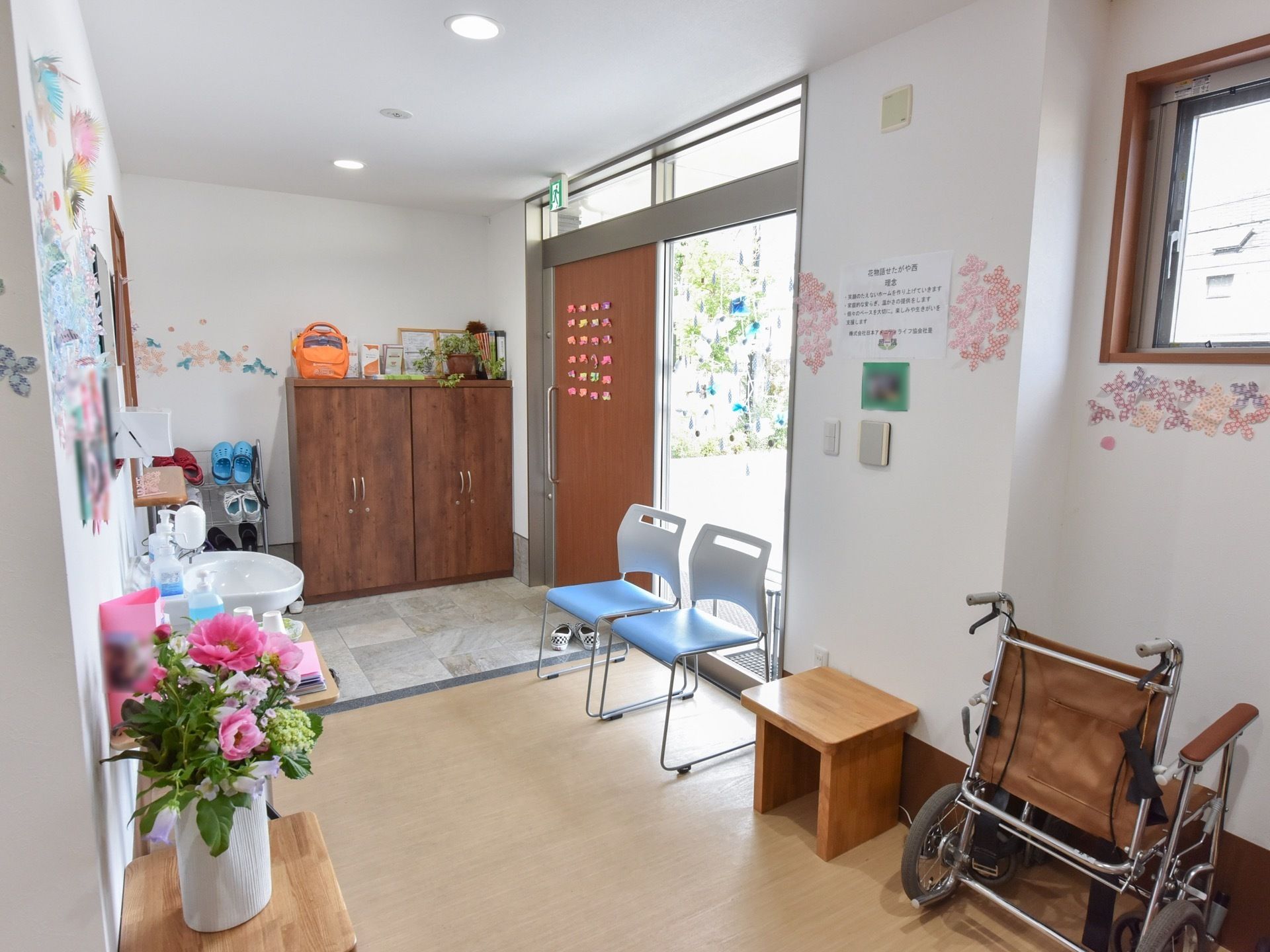 壁際に背の高い扉付きの茶色い靴箱が設置されている。青いパイプ椅子が２脚並べられ、車椅子が１台置いてある。