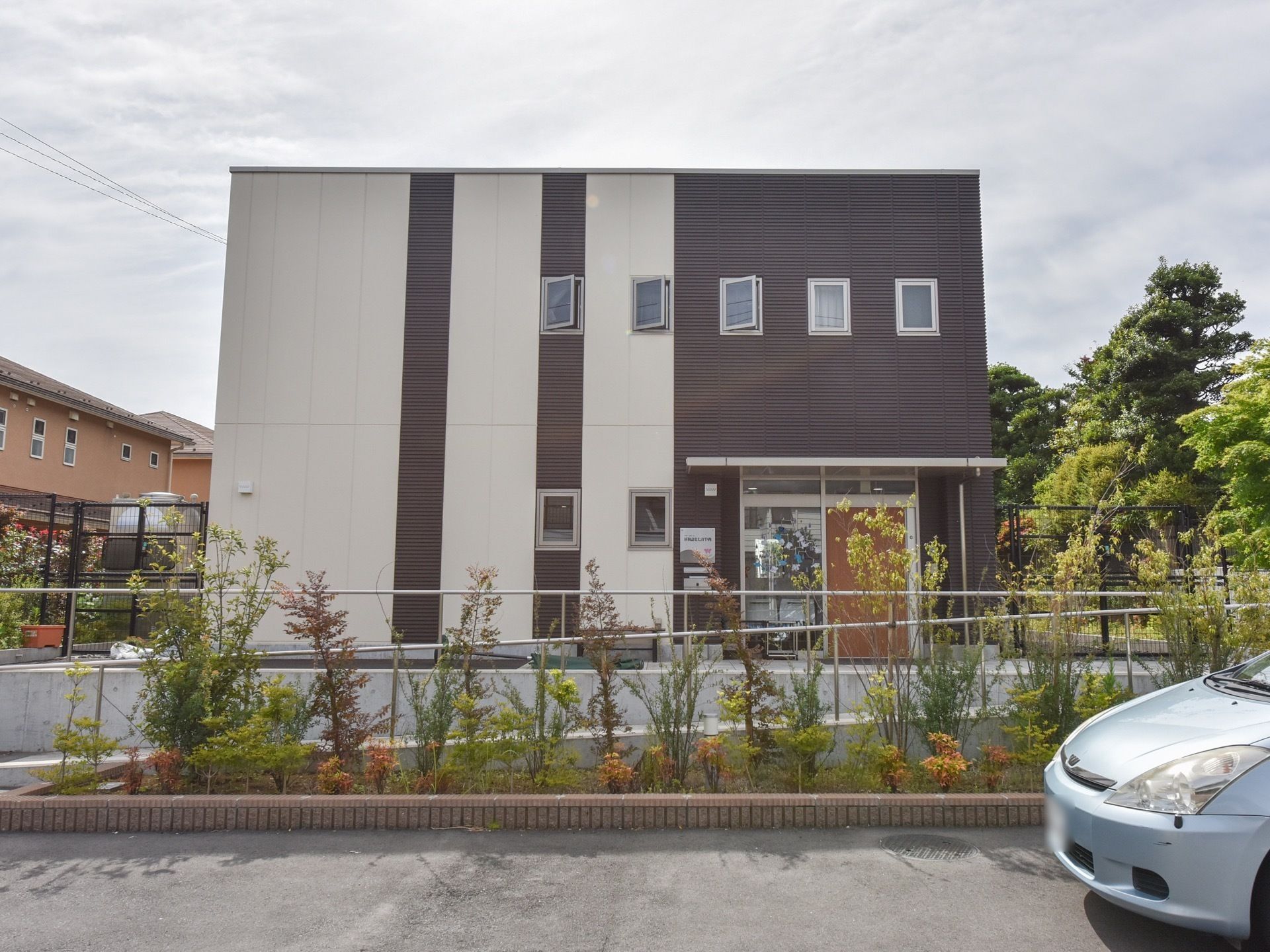 白と黒色の壁の２階建ての建物である。建物の周りにたくさんの樹木が植えられ、隣にオレンジ色の民家が建っている。