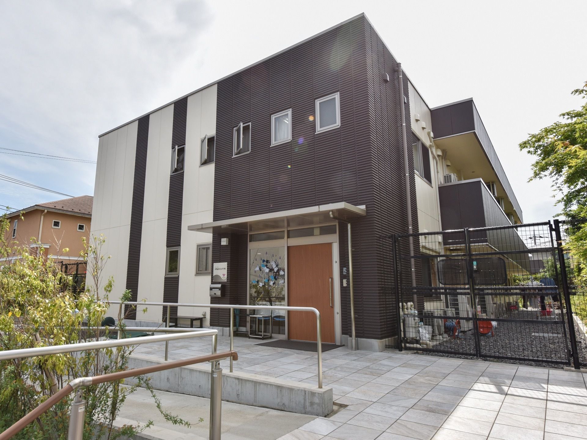 建物の敷地に砂利が敷かれ、黒いフェンスで仕切られている。２階に複数の小さな窓が取り付けられ、ベランダがある。
