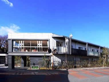白とグレーの落ちついた外壁の建物。１階部分は柵に沿って木が植えられ、２階部分にはベランダが設置されている。