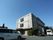 ブラウン調の３階建ての施設建物に、黒いサッシを使った出窓がついている。駐車スペースから玄関まではフラットで、点字シートが施工されている。