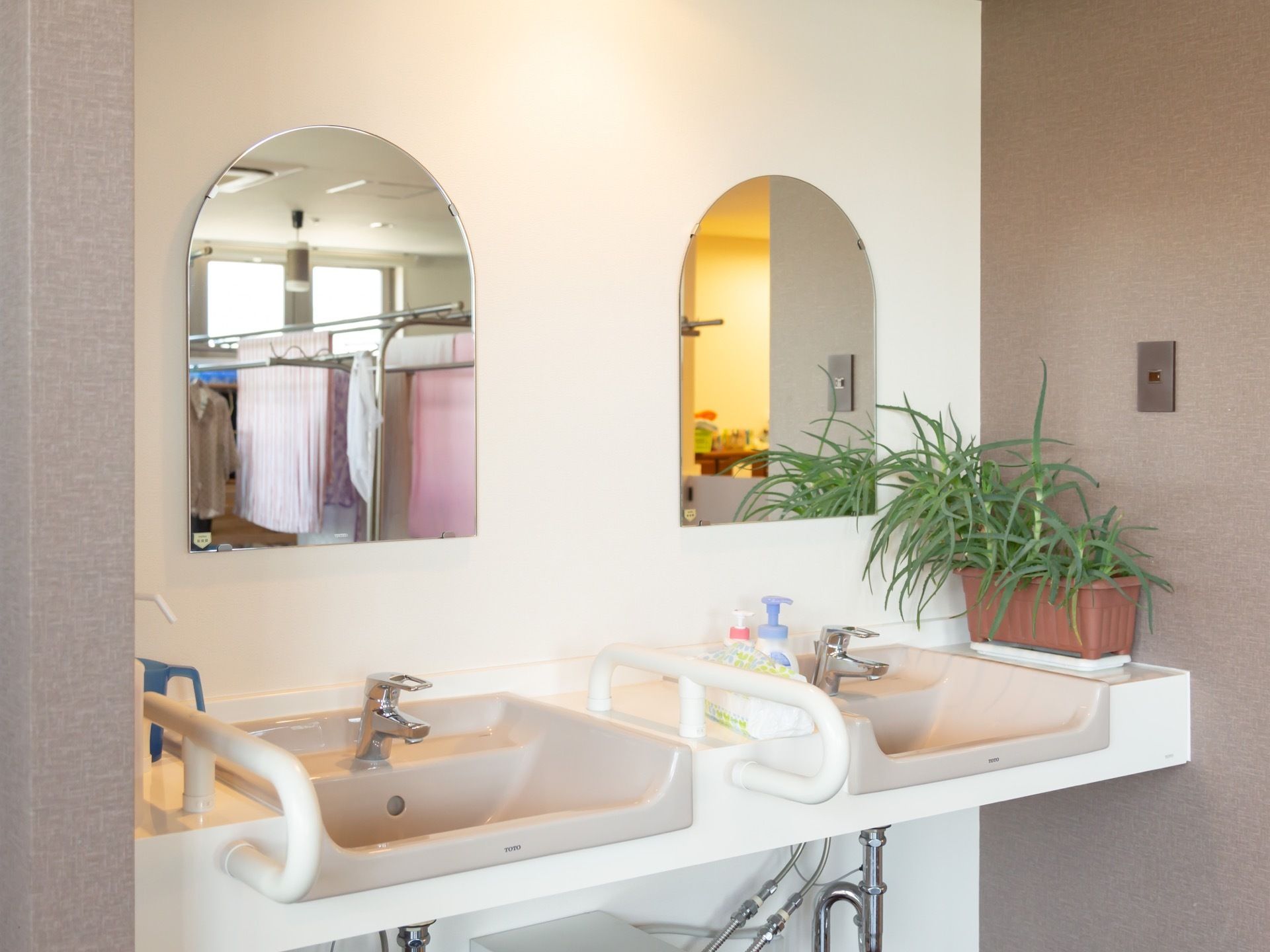 壁に2枚の鏡が取り付けられている洗面台。左側の洗面台には手すりがついている。白が基調でとても明るい雰囲気になっている。