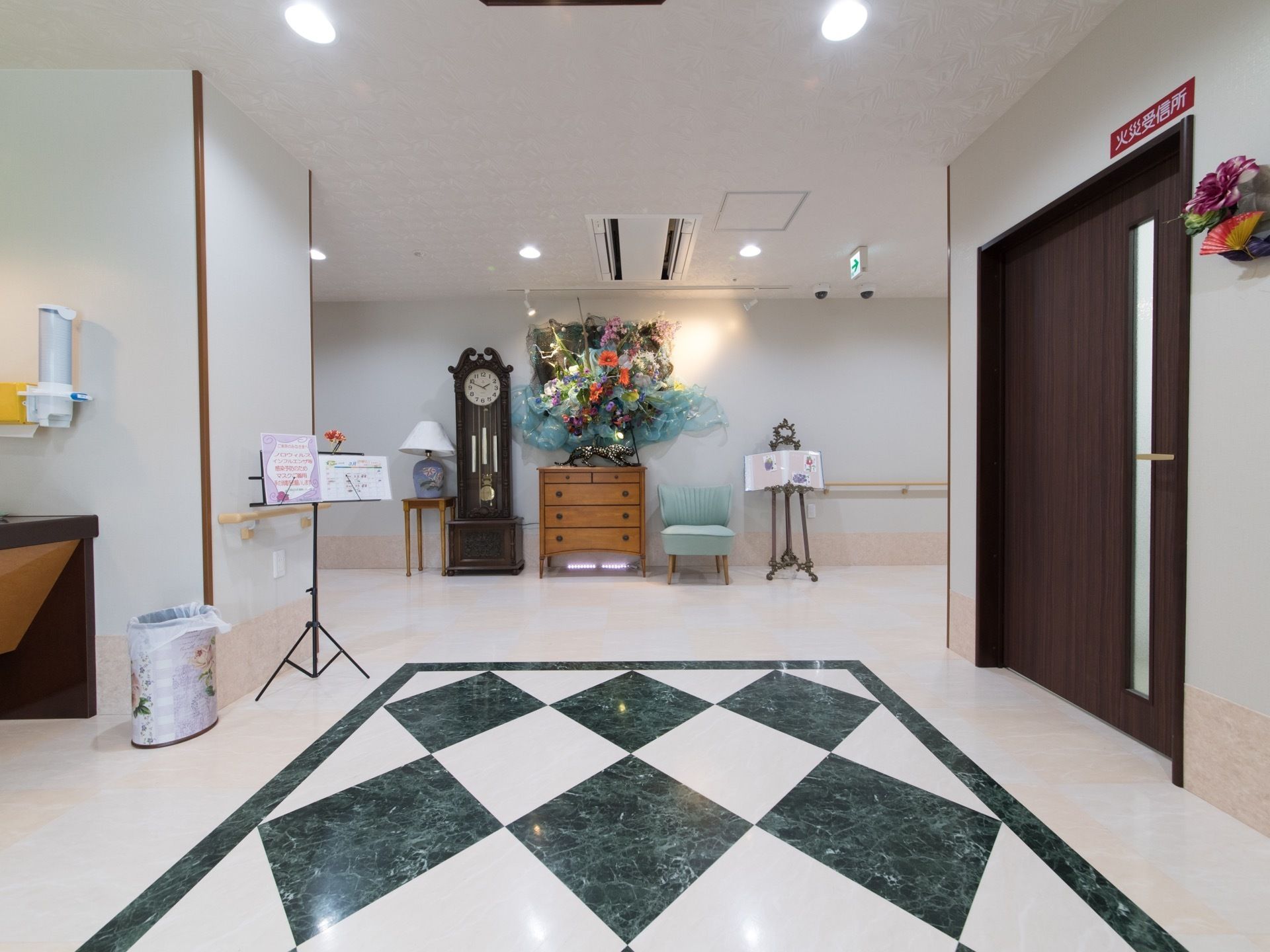 玄関内のつきあたりに、アンティーク風の時計やタンス、椅子が並べられている。右には火災受診所の扉がある。