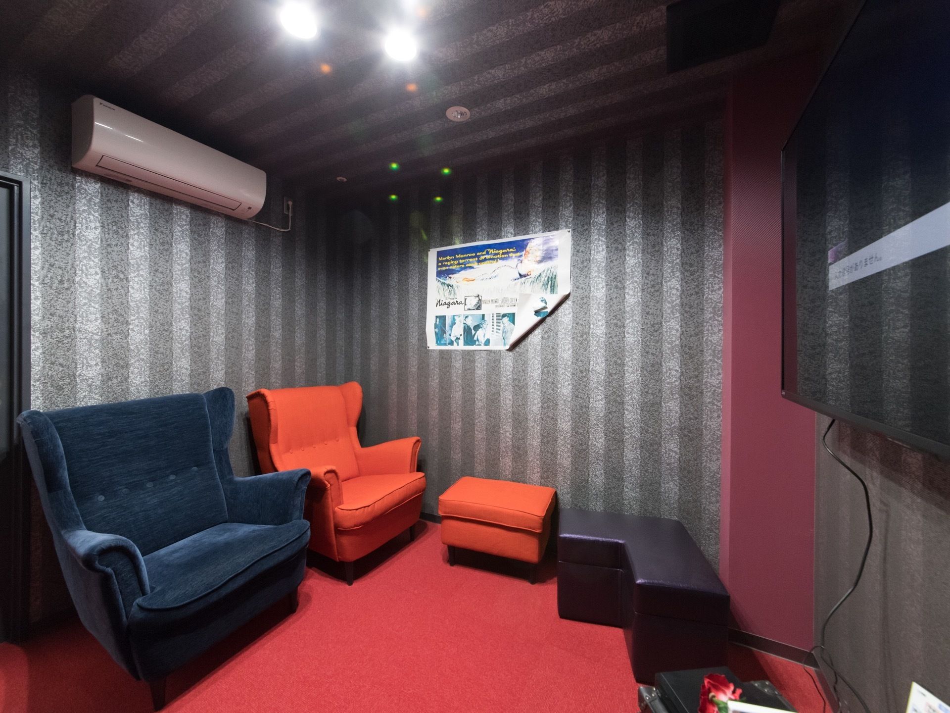 ストライプの壁に彩られた部屋の壁に、大きなテレビが設置されている。1人用の黒と赤のソファ、ベンチ型の椅子が置かれている。