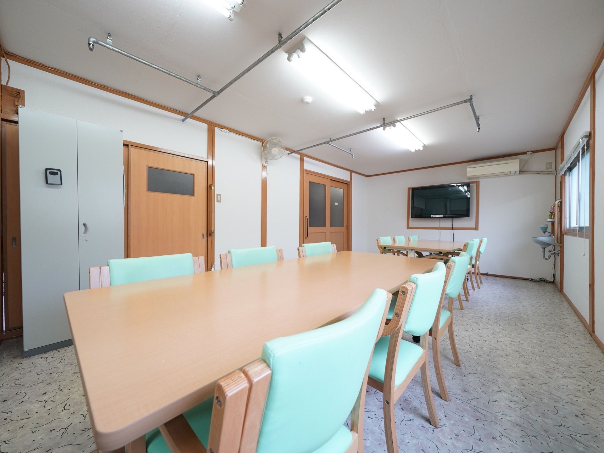 ６人掛けのテーブルが二台並んで置かれている。壁には大型のテレビが掛けられており、洗面台も設置されている。