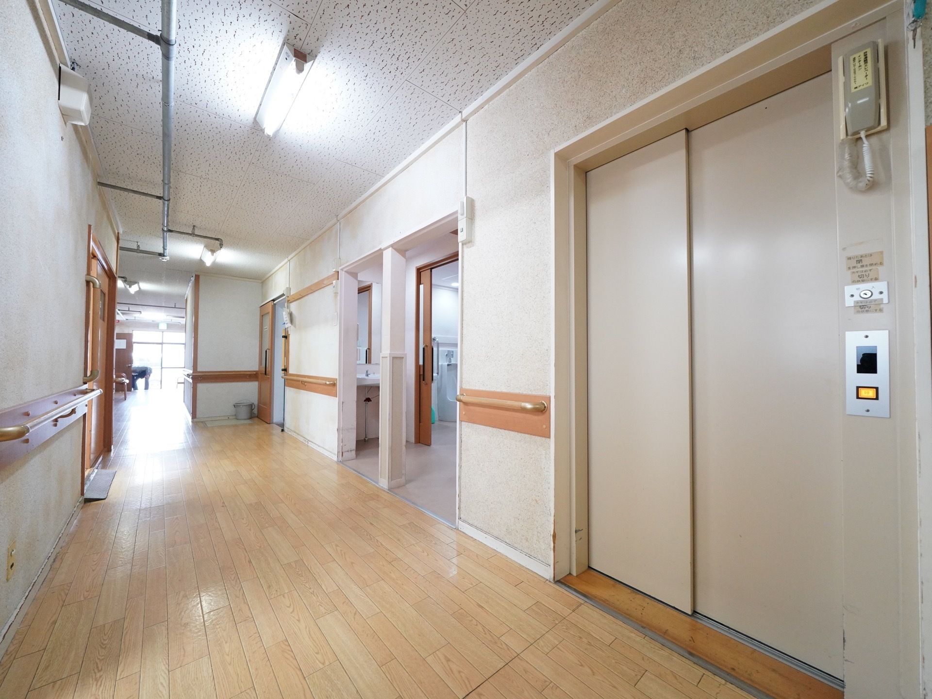 フローリングの床に、模様の入った白色の壁の廊下である。壁には手すりが設置されており、右手前にエレベーターが備わっている。
