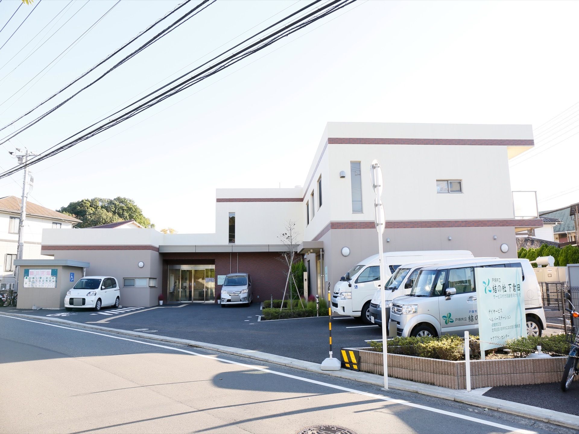 施設の建物の手前敷地内に、駐車スペースが設けられている。角には植栽された花壇があり、看板が立てられている。