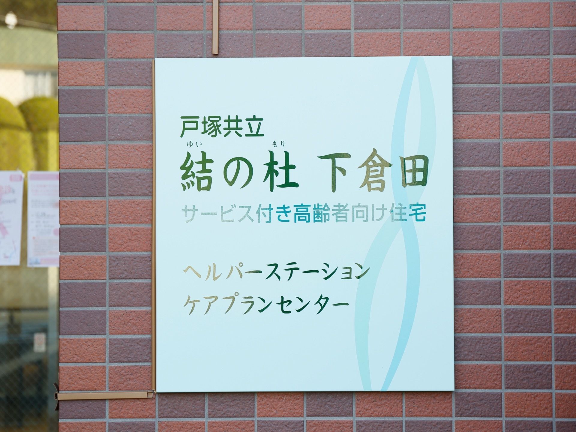 玄関の入り口周りのレンガの壁に、施設の名前を記した看板がかかっている。ふりがなが振られている。薄いブルーの看板である。