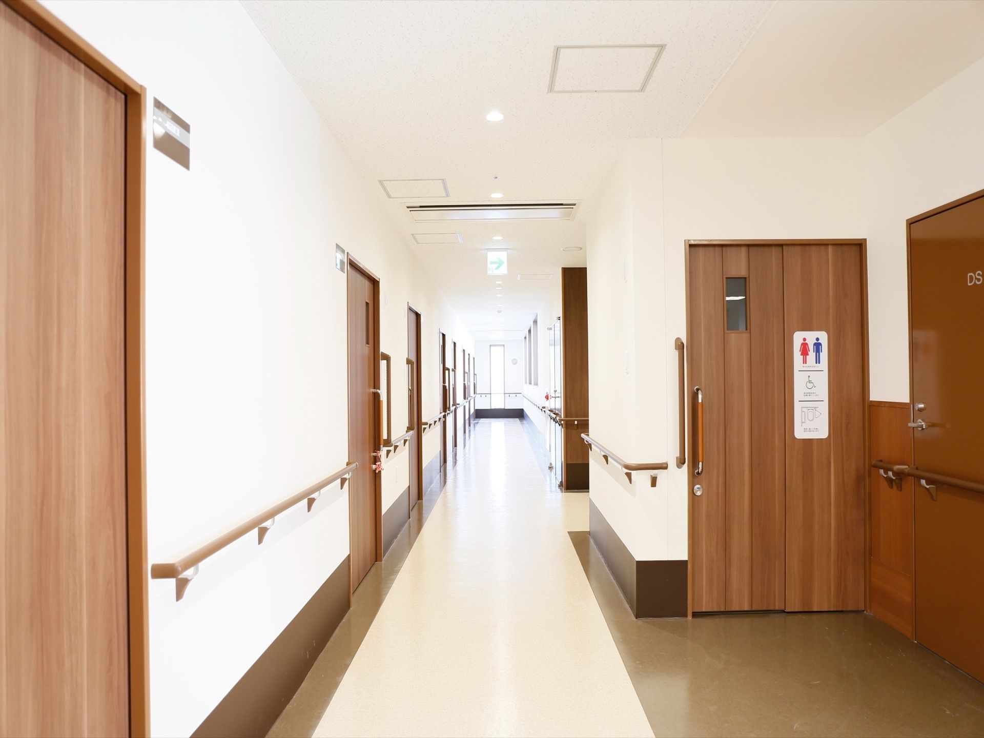 廊下の左側に、居室の扉が並んでる。右側に多目的トイレがある。扉はすべてダークブラウンの木目のものである。