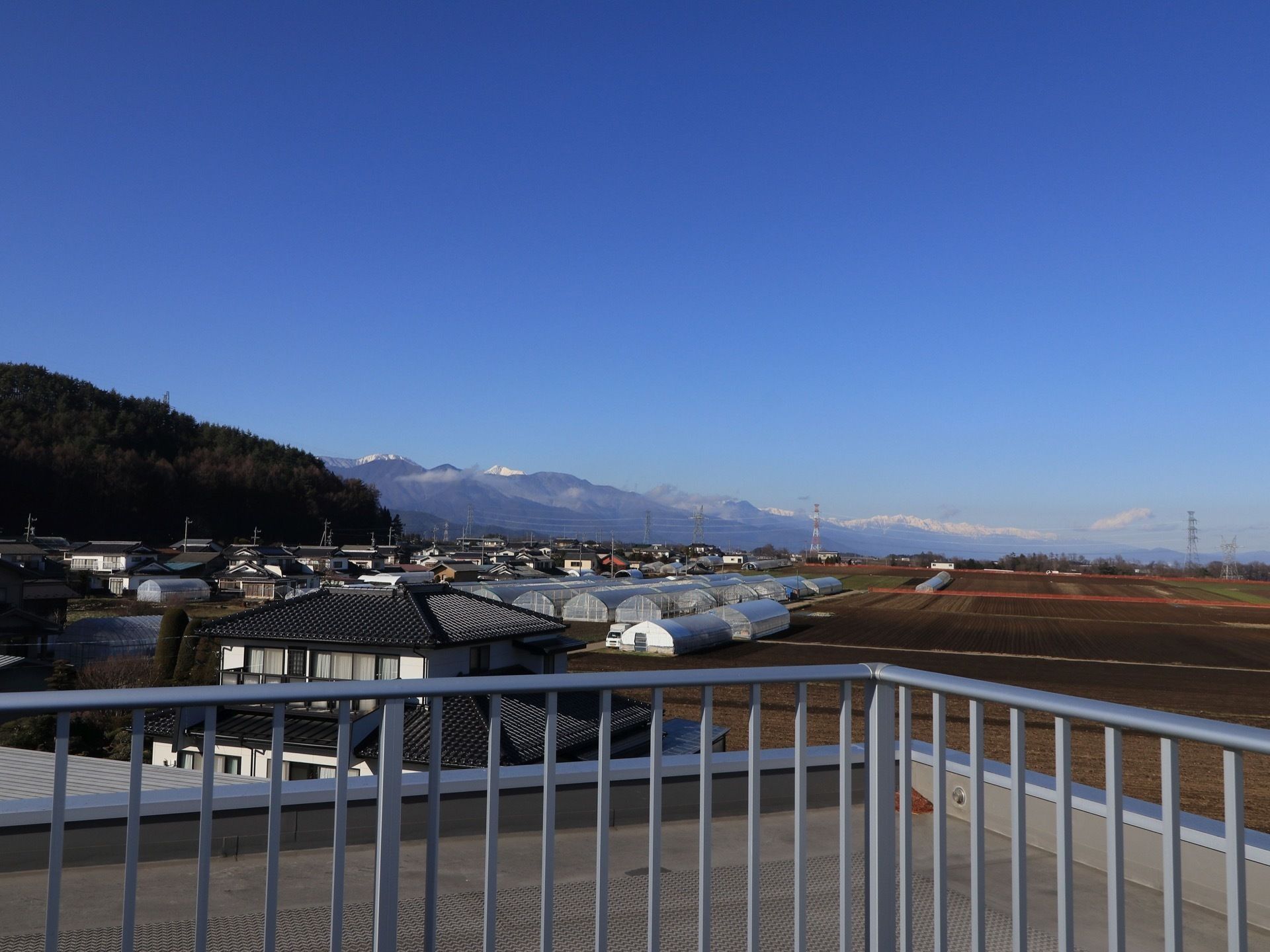 建物の屋上からの風景である。民家やビニールハウス、畑、緑豊かな山を望むことができる。転落防止の柵が設置されている。