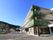 建物の角部分に描かれた大きな木が特徴的な３階建て施設である。広々とした駐車場が建物前に設けられている。