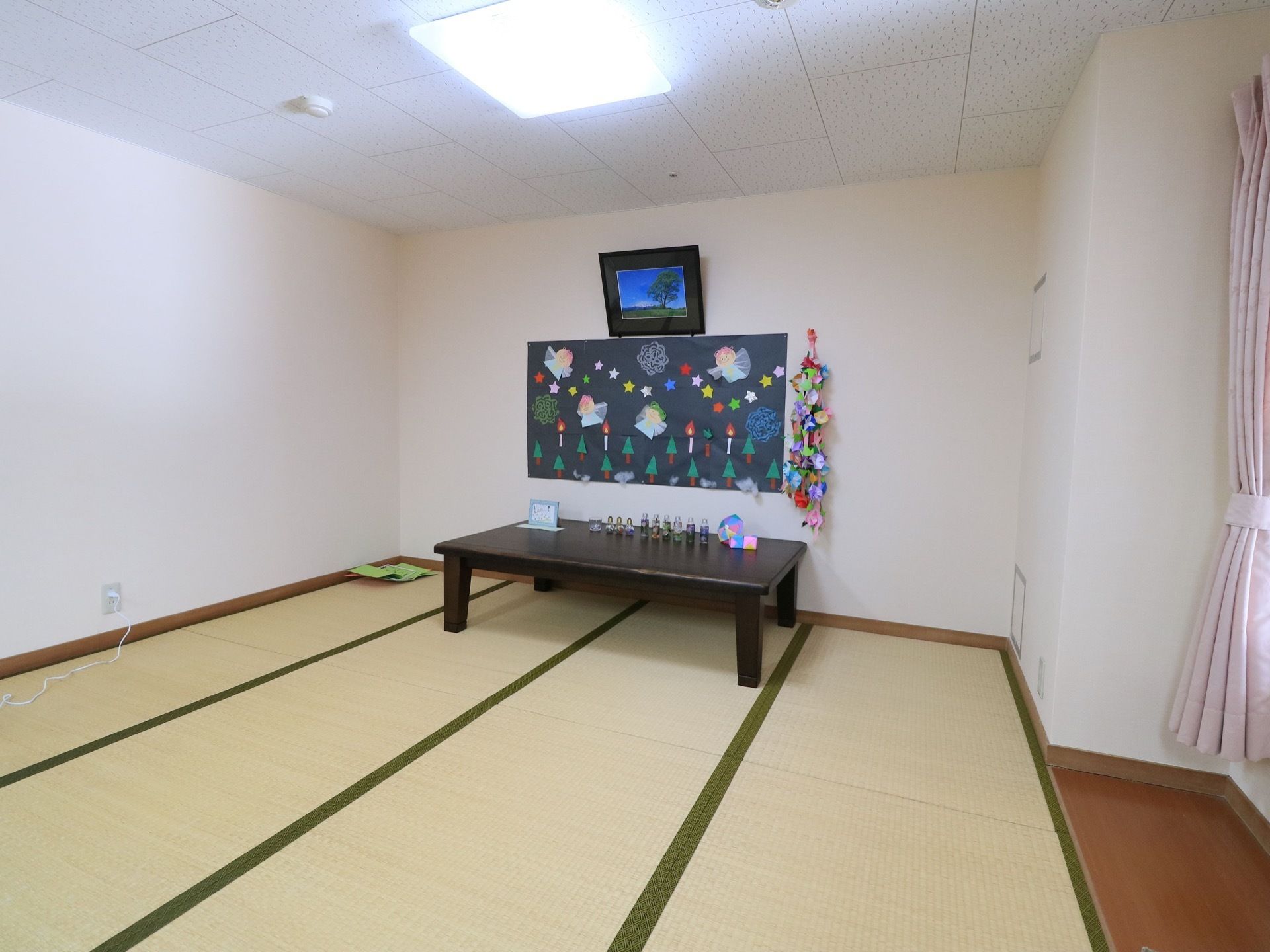 畳敷きの部屋に、テーブルが置かれている。その上にこけしや人形などが並べられている。壁にはロウソクや木を描いた絵が貼られている。