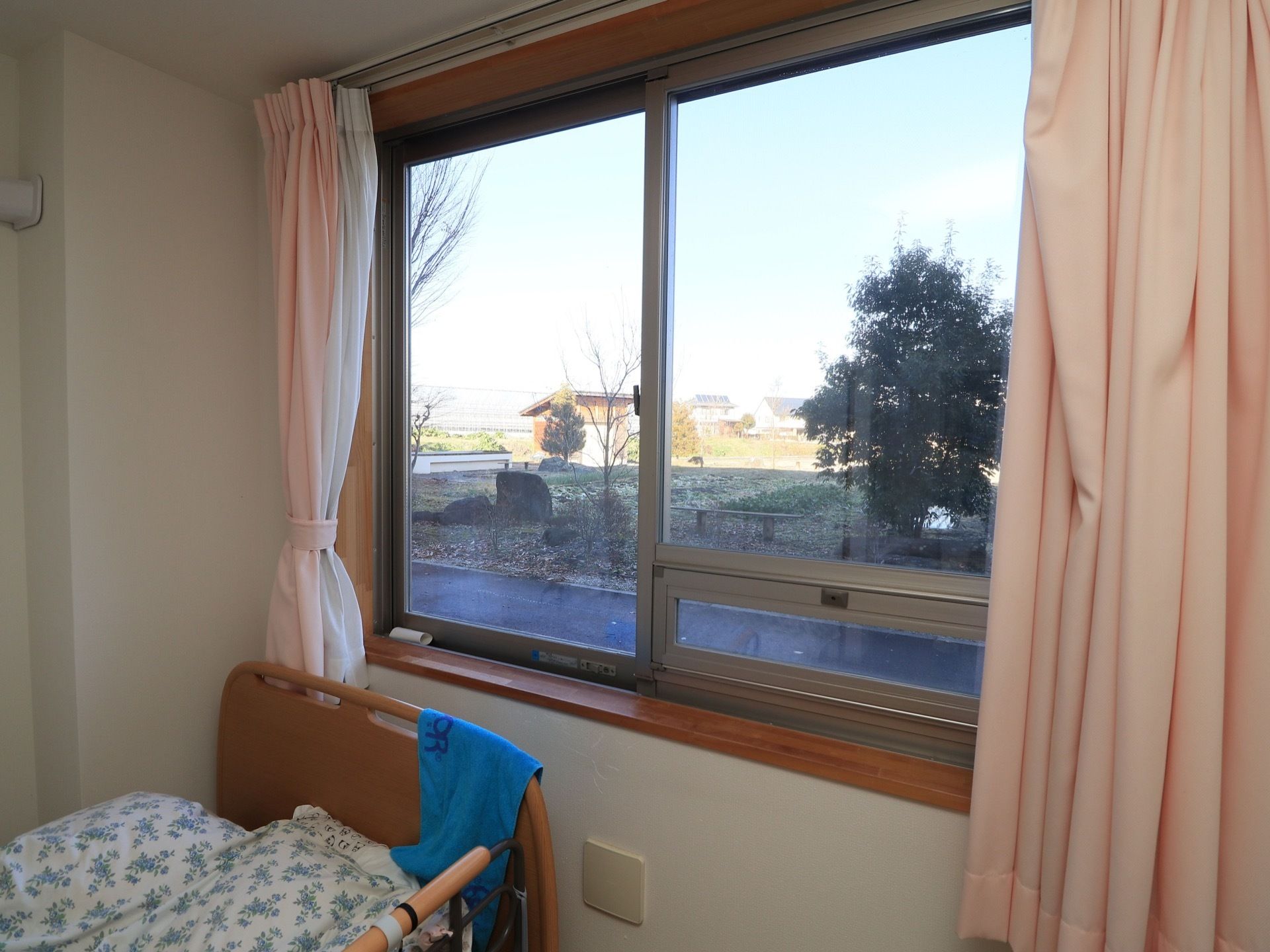居室には、腰の高さほどの場所に窓がある。ピンク色のカーテンがかかっている。窓際にはベッドが置かれている。