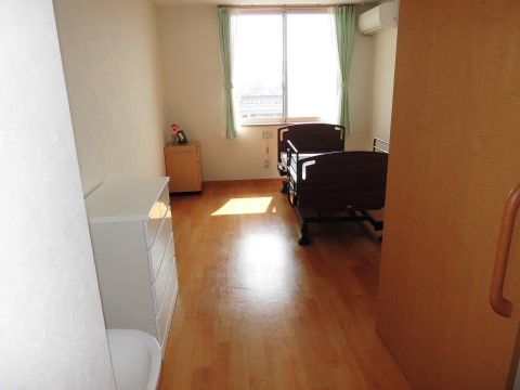 窓から明るい光の差す居室にエアコンが設置されている。手すり付きベッドの枕元にはナースコールボタンが設置されている。