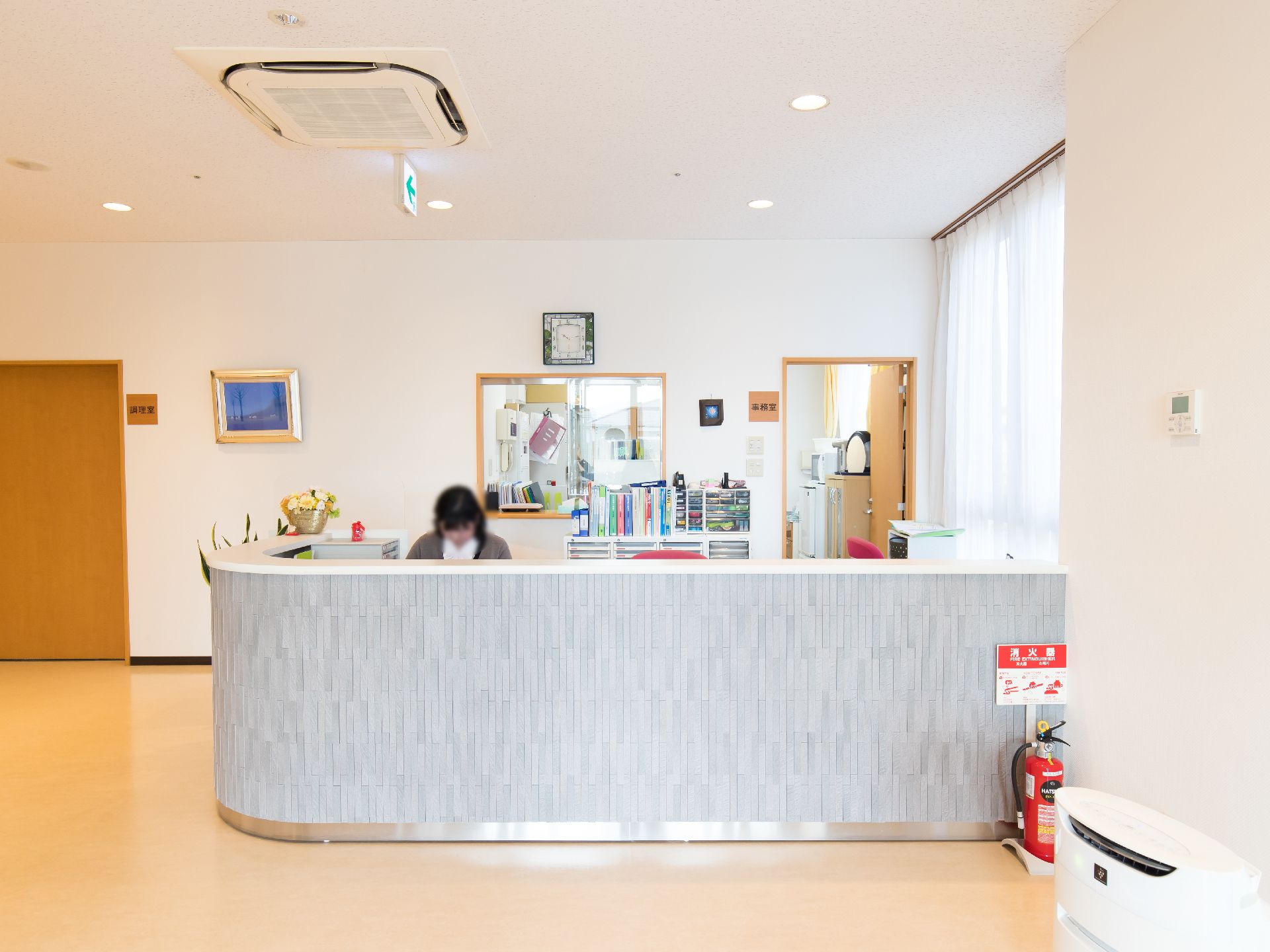 グレーの受付カウンターに女性スタッフが１人座っている。奥に部屋があり、扉の左側に事務室と書かれている。