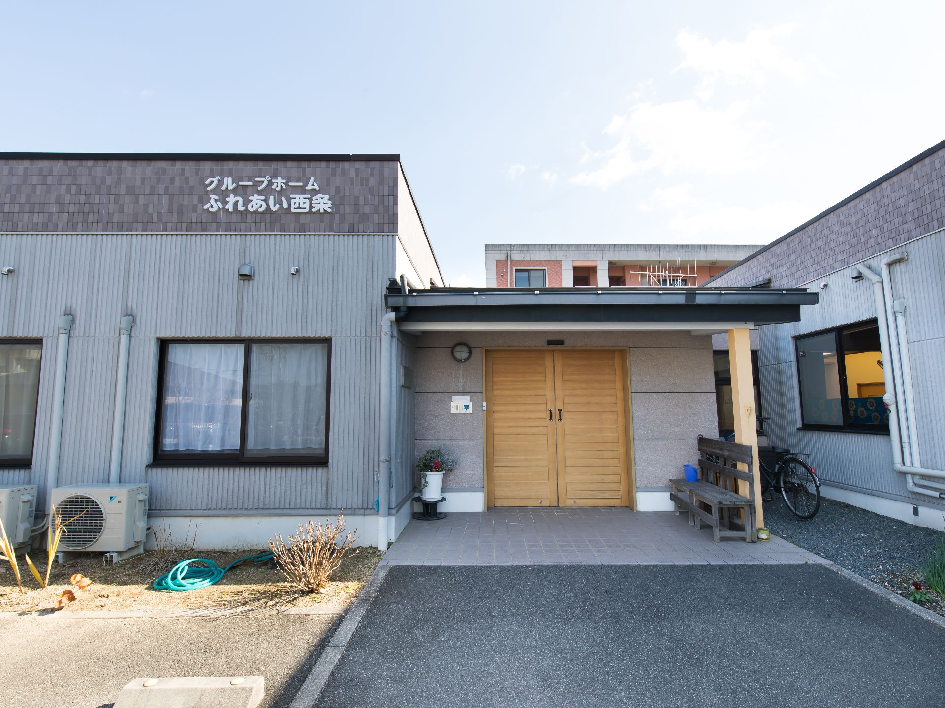 庇の下にベンチとプランターが飾られている、両開き扉の平屋建て