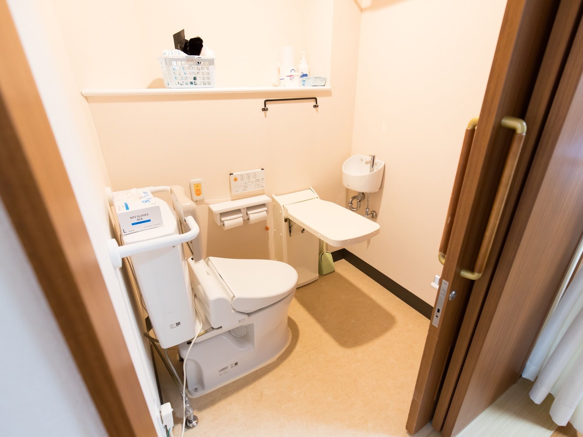 跳ね上げ式アームレストや洗面台、温水洗浄便座が完備されたトイレである。便器後ろの壁には、背もたれが設置されている。