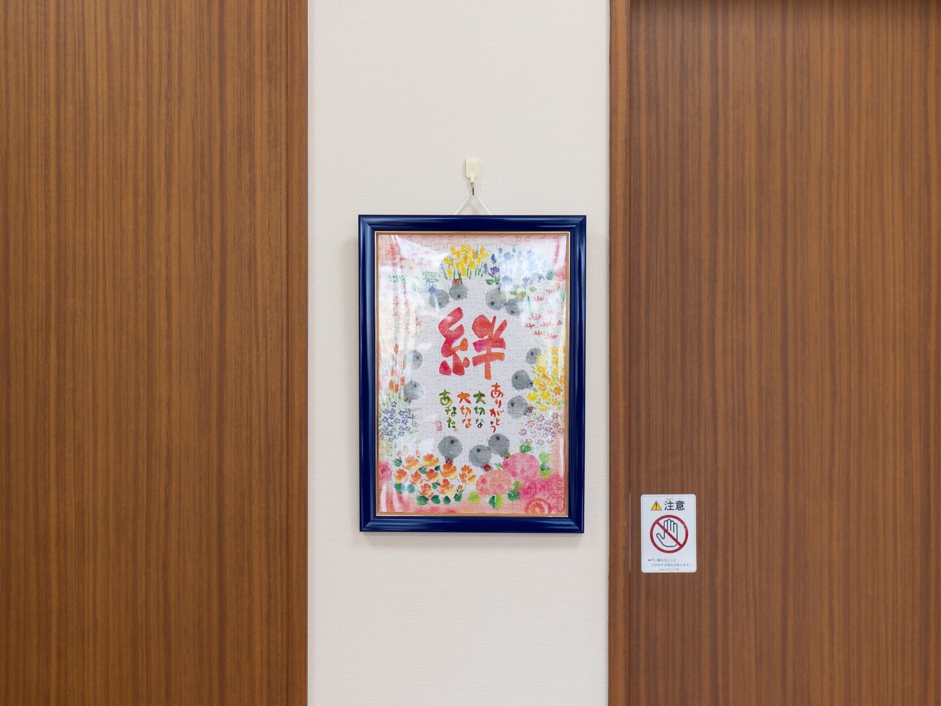 壁には、水彩絵の具で花や文字が描かれた絵が飾られている。右横には、扉が木目調のエレベーターが設置されている。