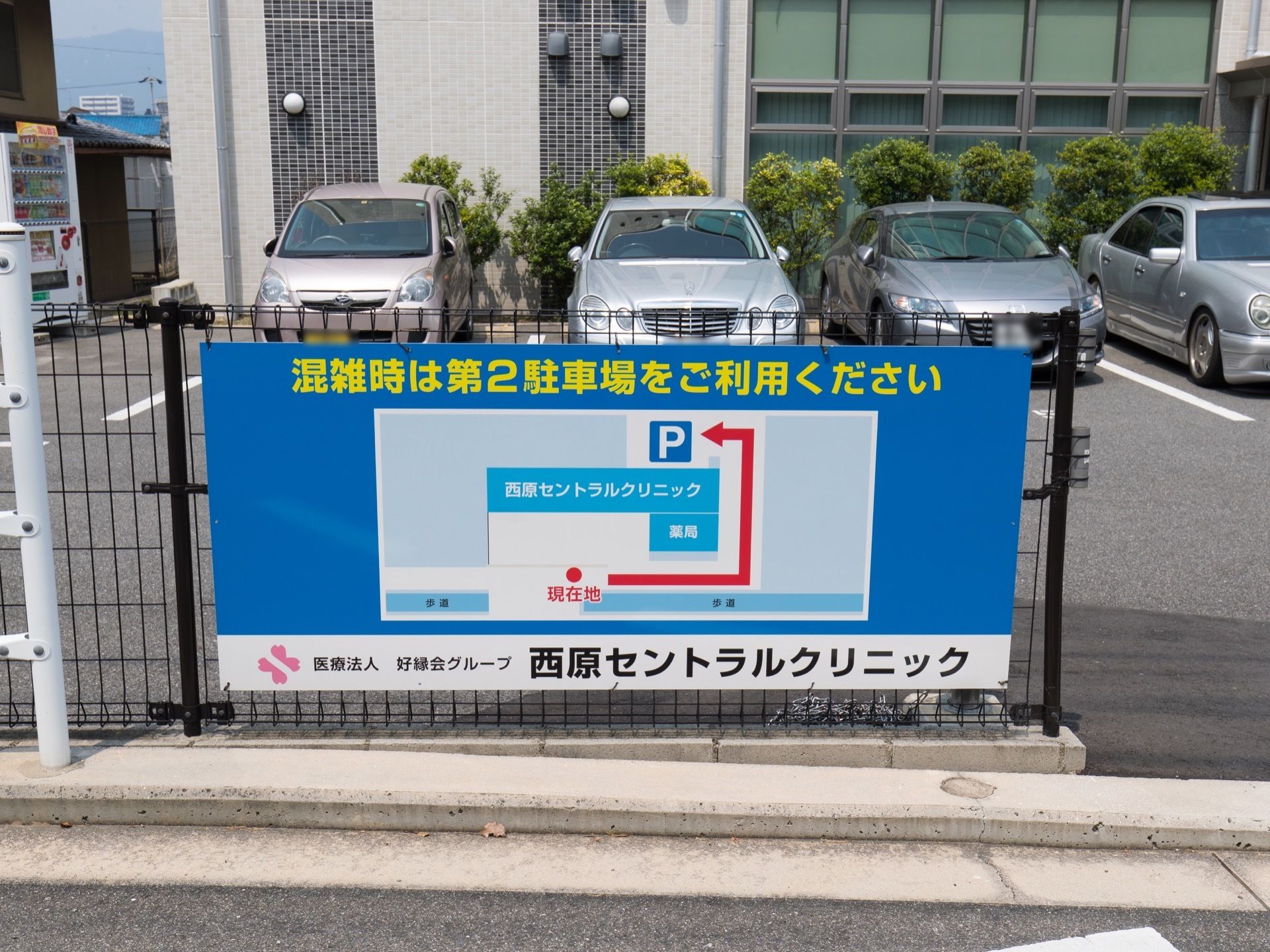 建物の前に駐車場があり、フェンスに第二駐車場の案内の看板がかかっている。クリニックと薬局を併設している。