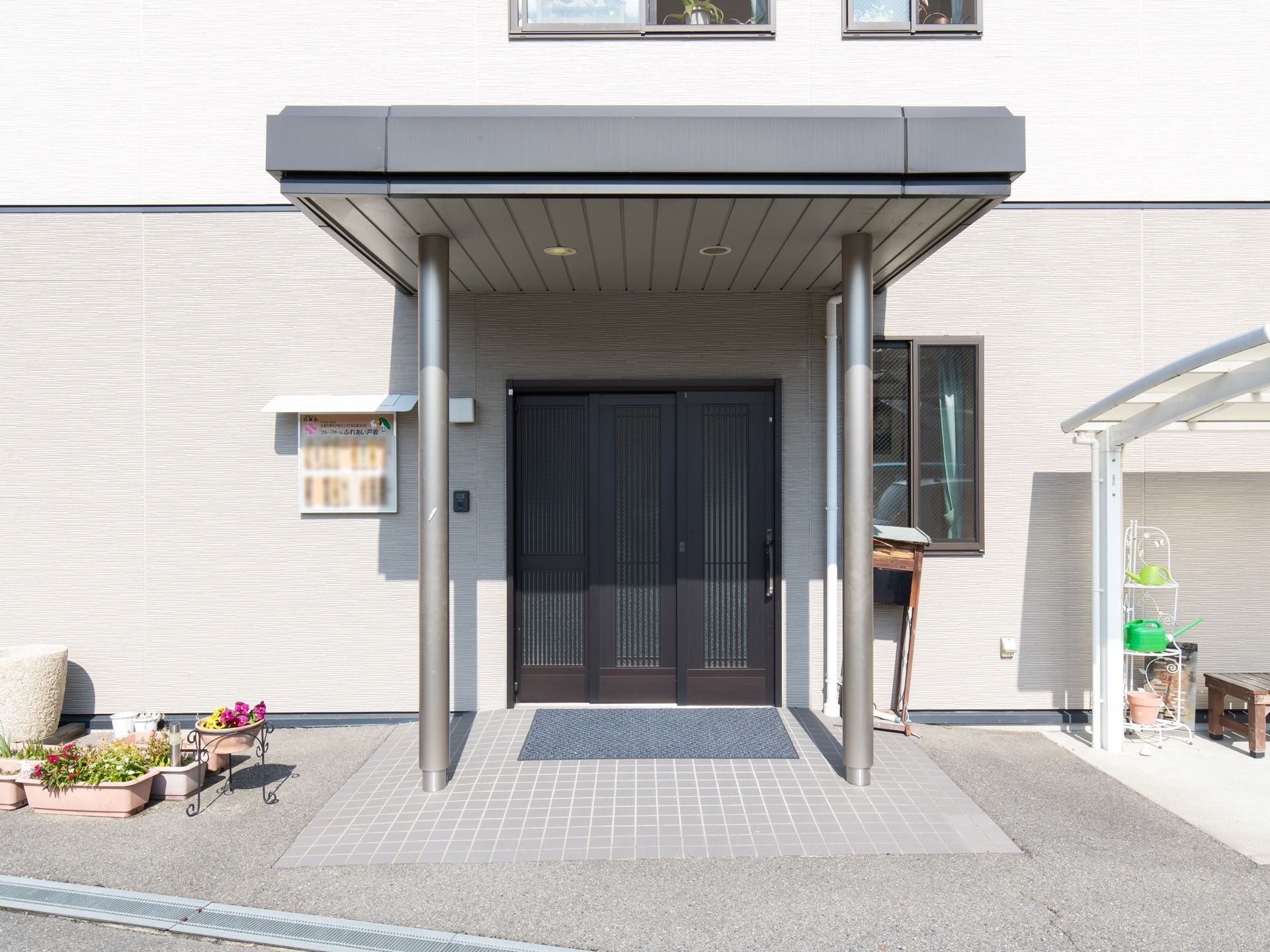 玄関前には屋根が設置されており、入口には黒い3枚引戸が取りつけられている。左側には、植木鉢に植物がたくさん植えられている。