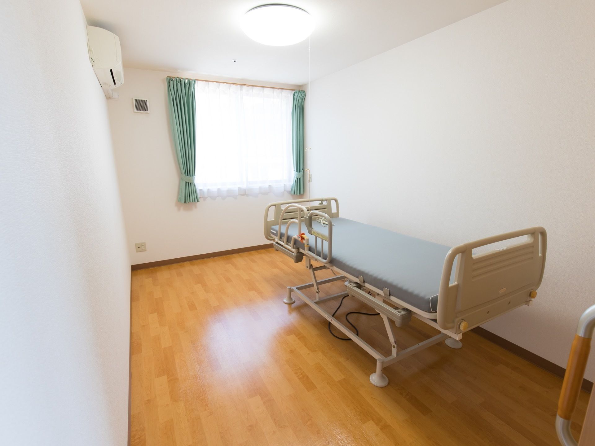エアコンが完備されている。窓から日が差し込んでいる。介護用ベッドが置かれている。段差のないフローリングである。