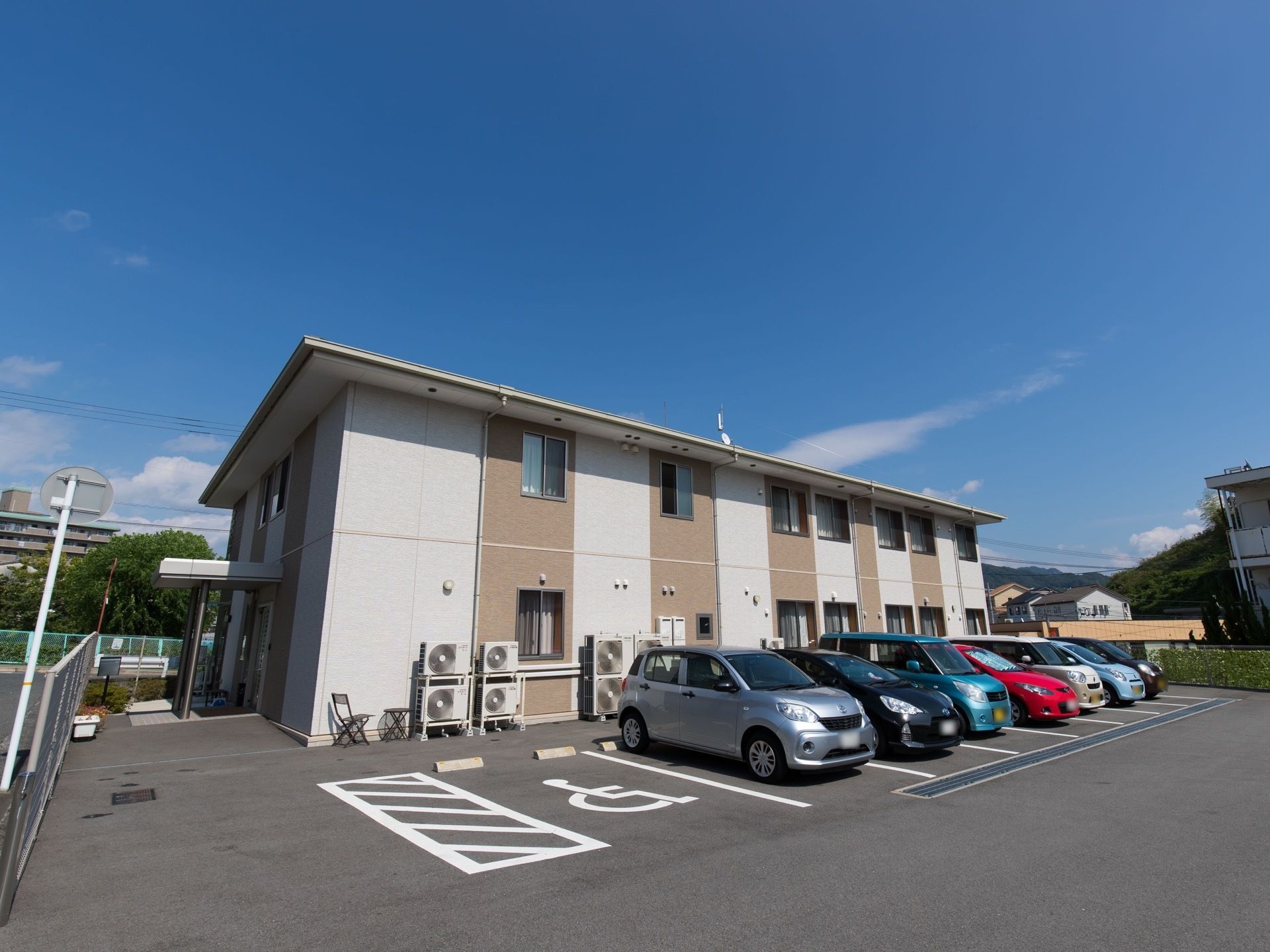 施設は集合住宅のような外観の建物である。駐車スペースが建物横に設けられている。壁際に椅子とテーブルが置かれている。