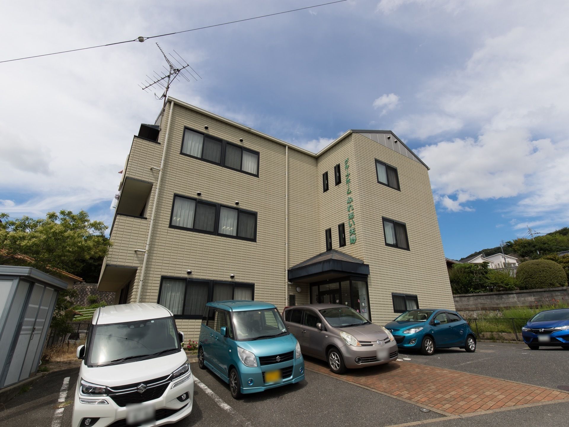 周囲に緑があり目の前が駐車場になっている、ベランダ付の建物