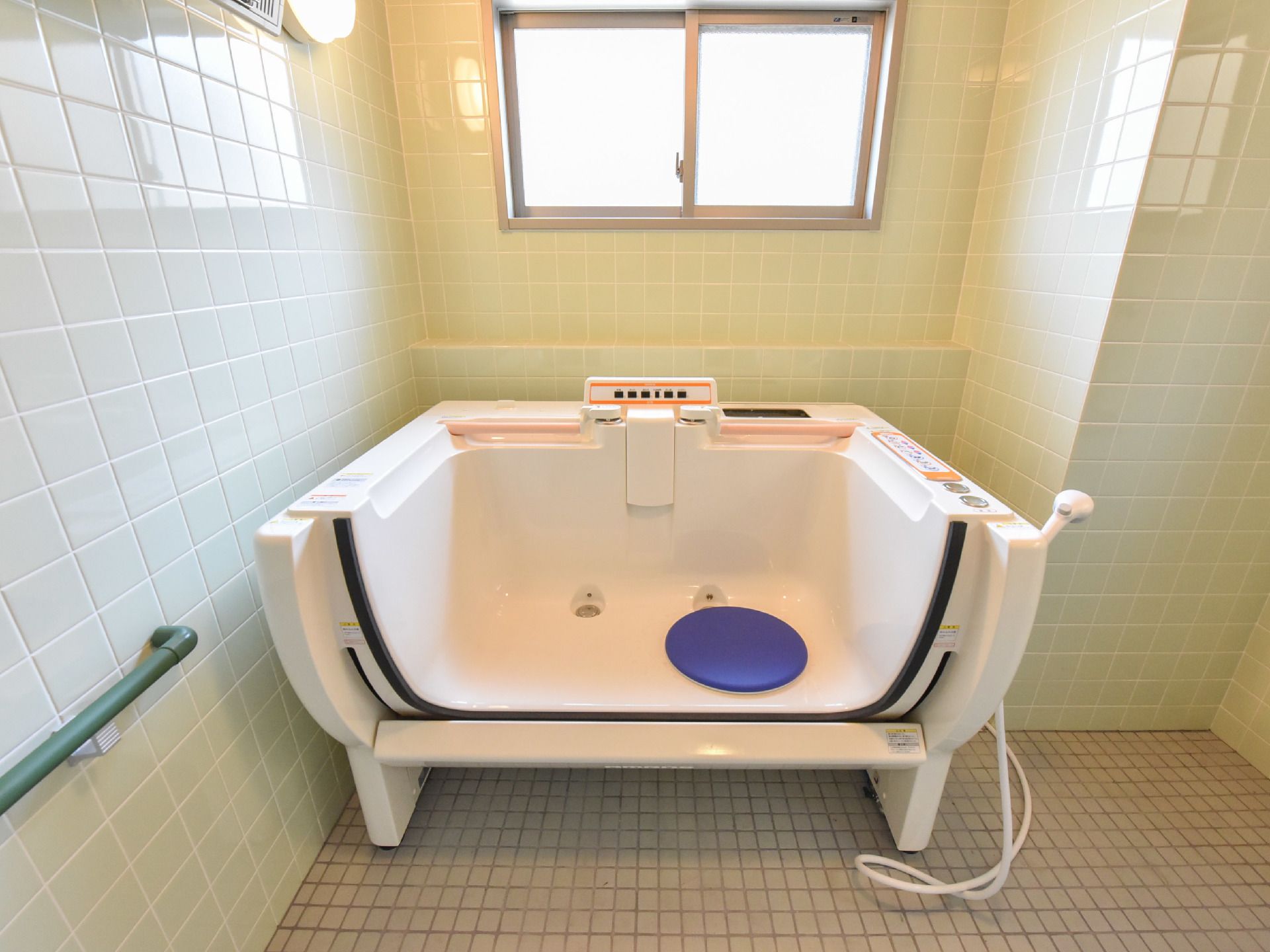 正面には介護入浴装置があり、その上側には窓が付いている。左手前には手すりが付いている。左上には照明器具が付いている。
